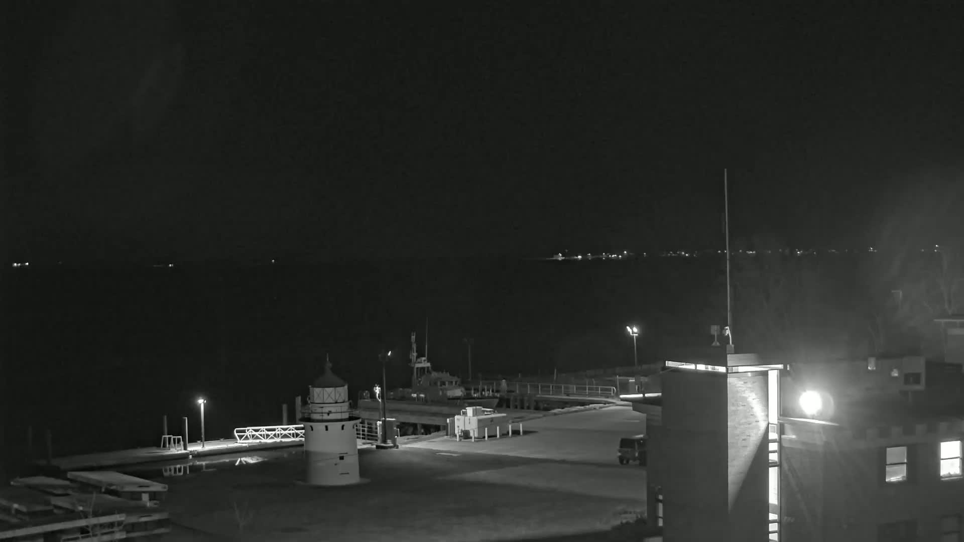 A nighttime, black and white view of a harbor with a lighthouse, a small boat, and a building, under a dark sky.