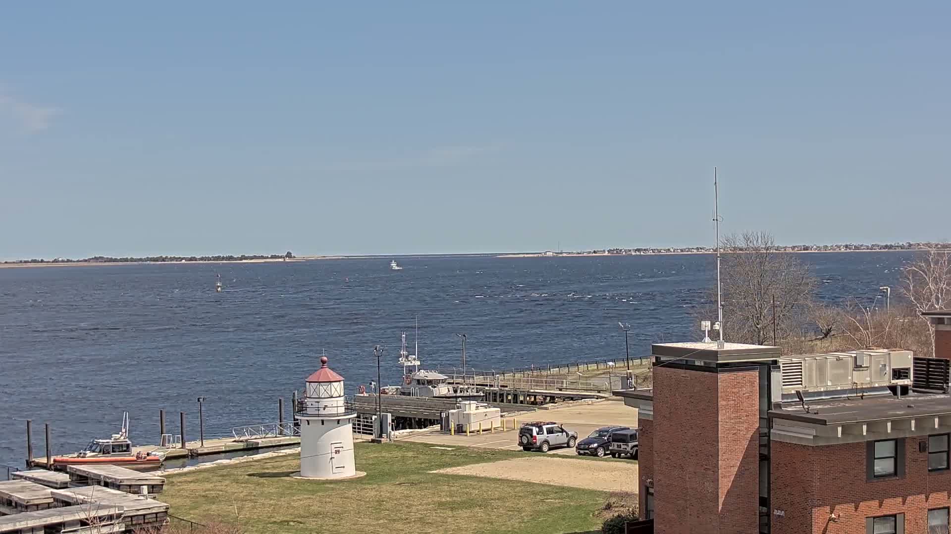 A small lighthouse and a docked boat are visible near a brick building overlooking a choppy body of water under a clear blue sky.