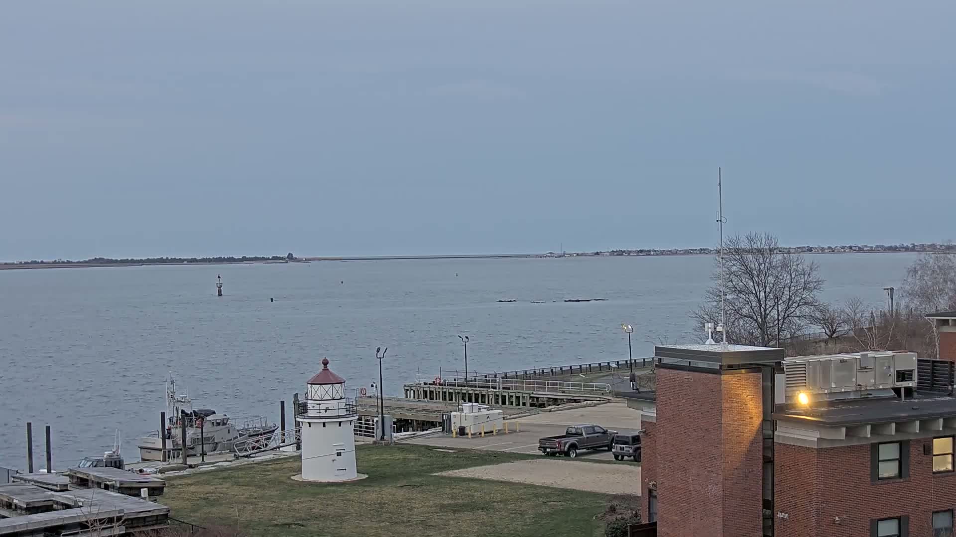 A small lighthouse sits on a grassy area next to a harbor with a boat and a dock, under an overcast sky.