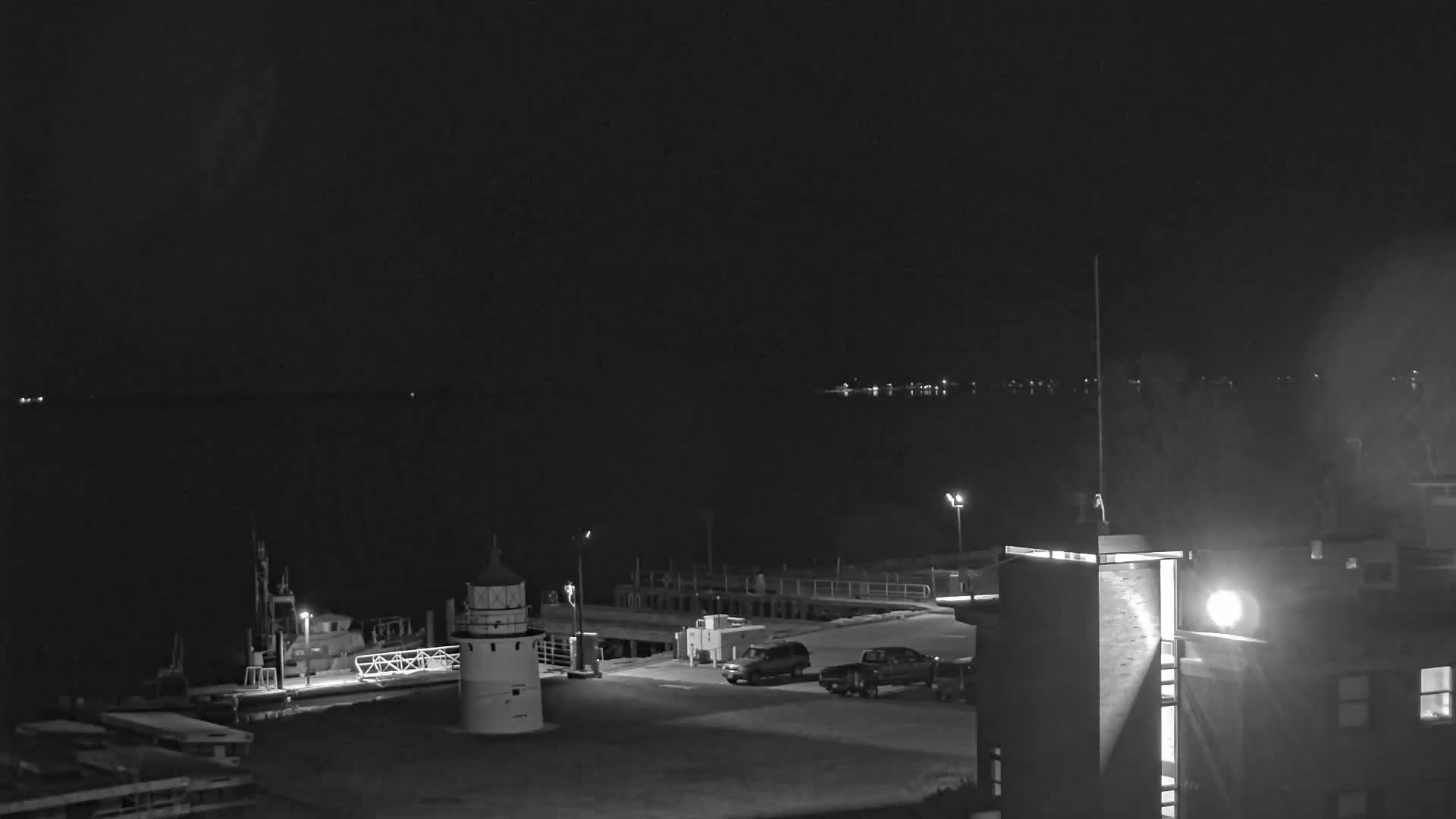 A nighttime view of a harbor with a lighthouse, boats, and vehicles parked near a building, under a clear, dark sky.