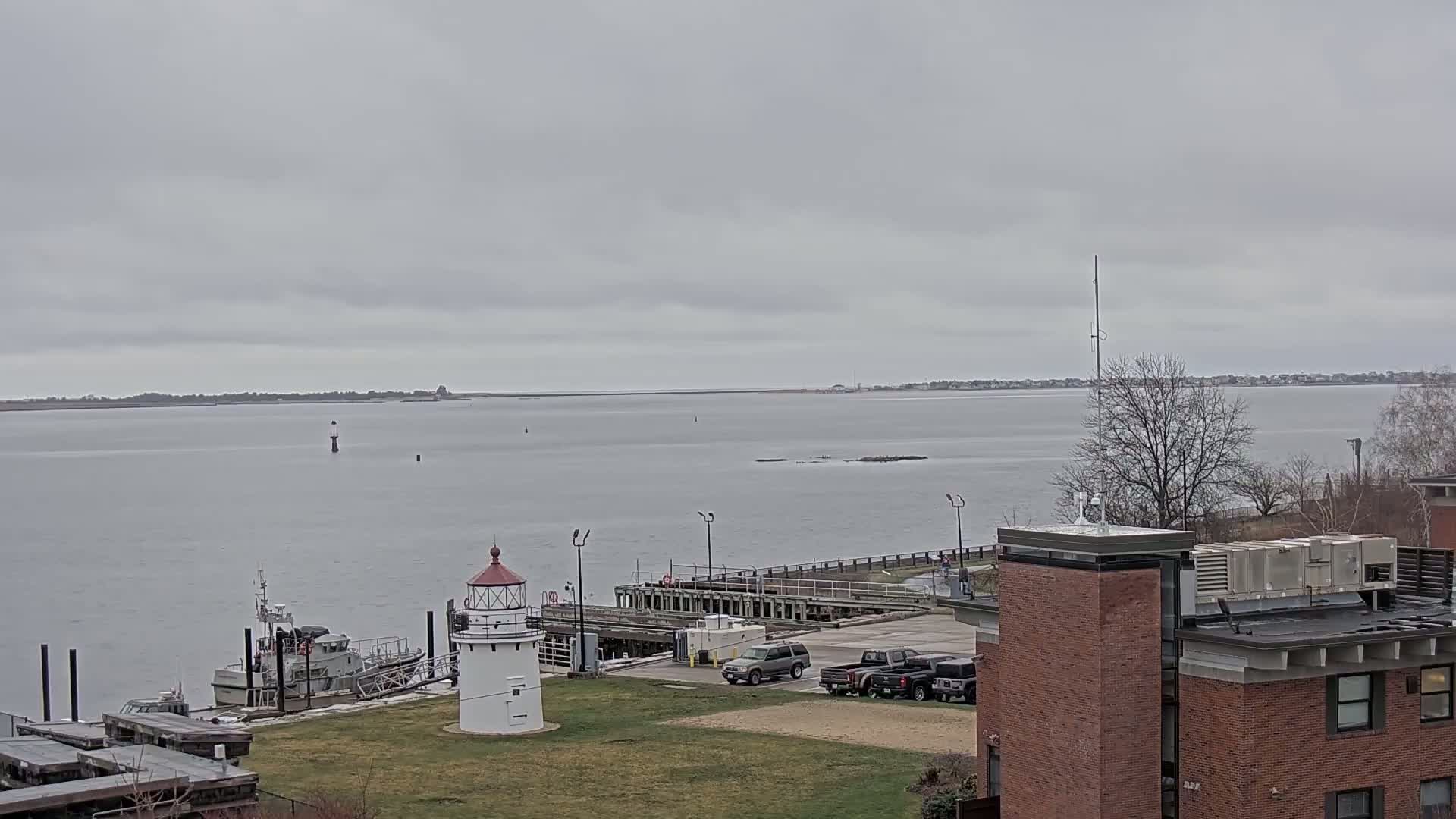 A small white lighthouse stands on a grassy area near a harbor, with a Coast Guard vessel moored nearby, under an overcast sky.