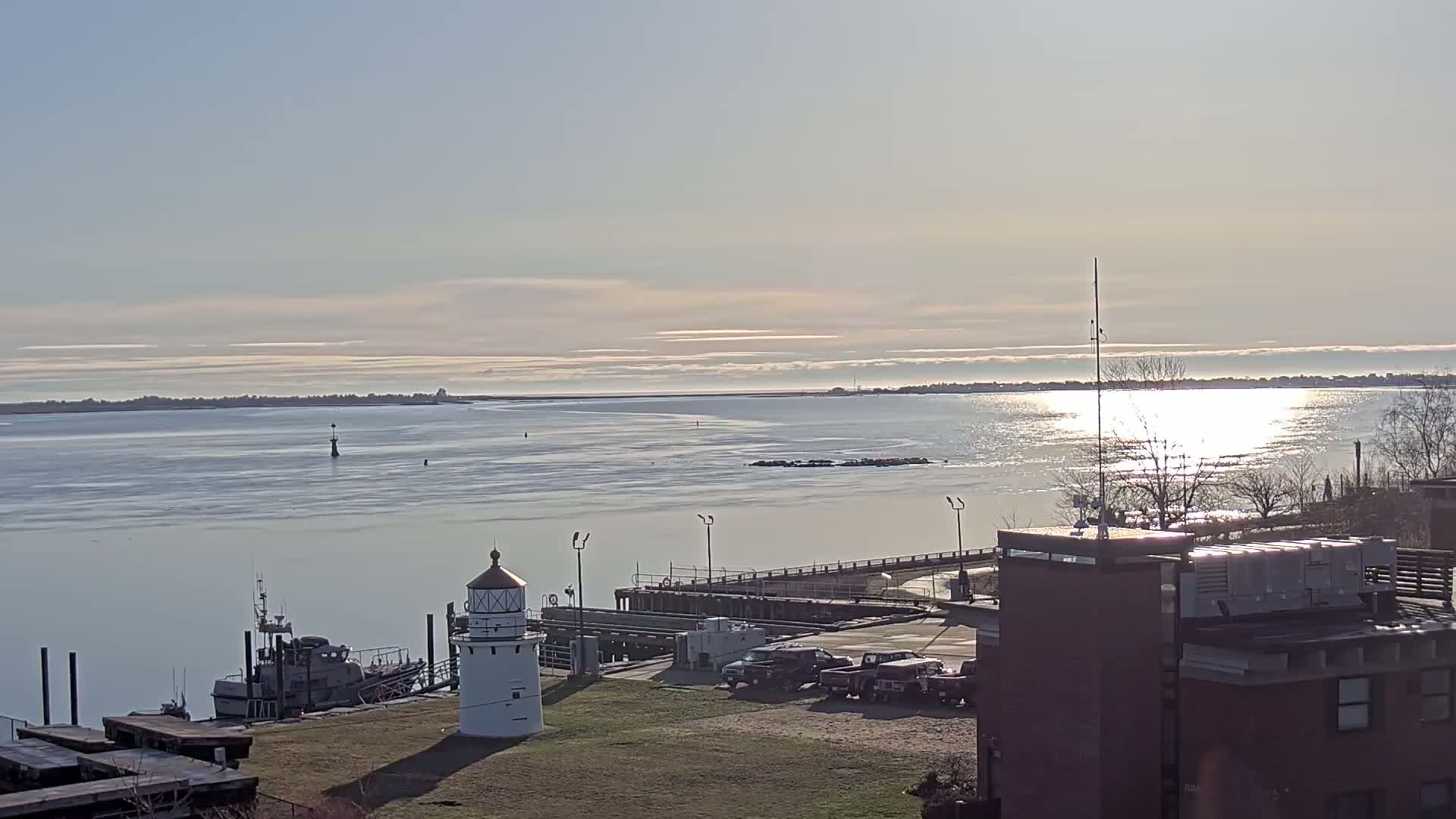 A small lighthouse stands near a harbor, with a calm body of water reflecting sunlight under a mostly clear sky; a small boat is docked nearby.