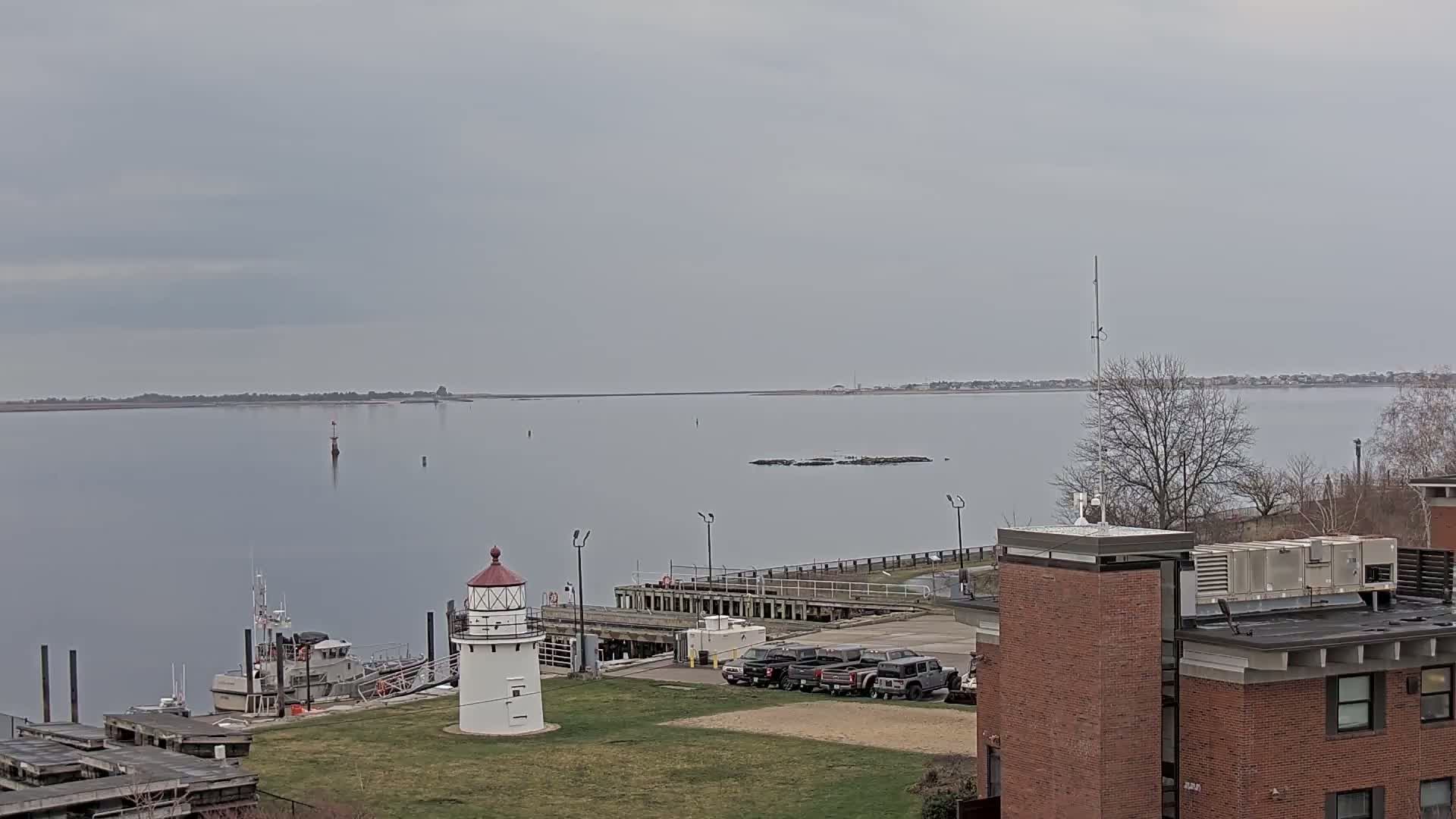 A small lighthouse sits on a grassy area next to a dock with several vehicles and a boat, overlooking a calm body of water under an overcast sky.