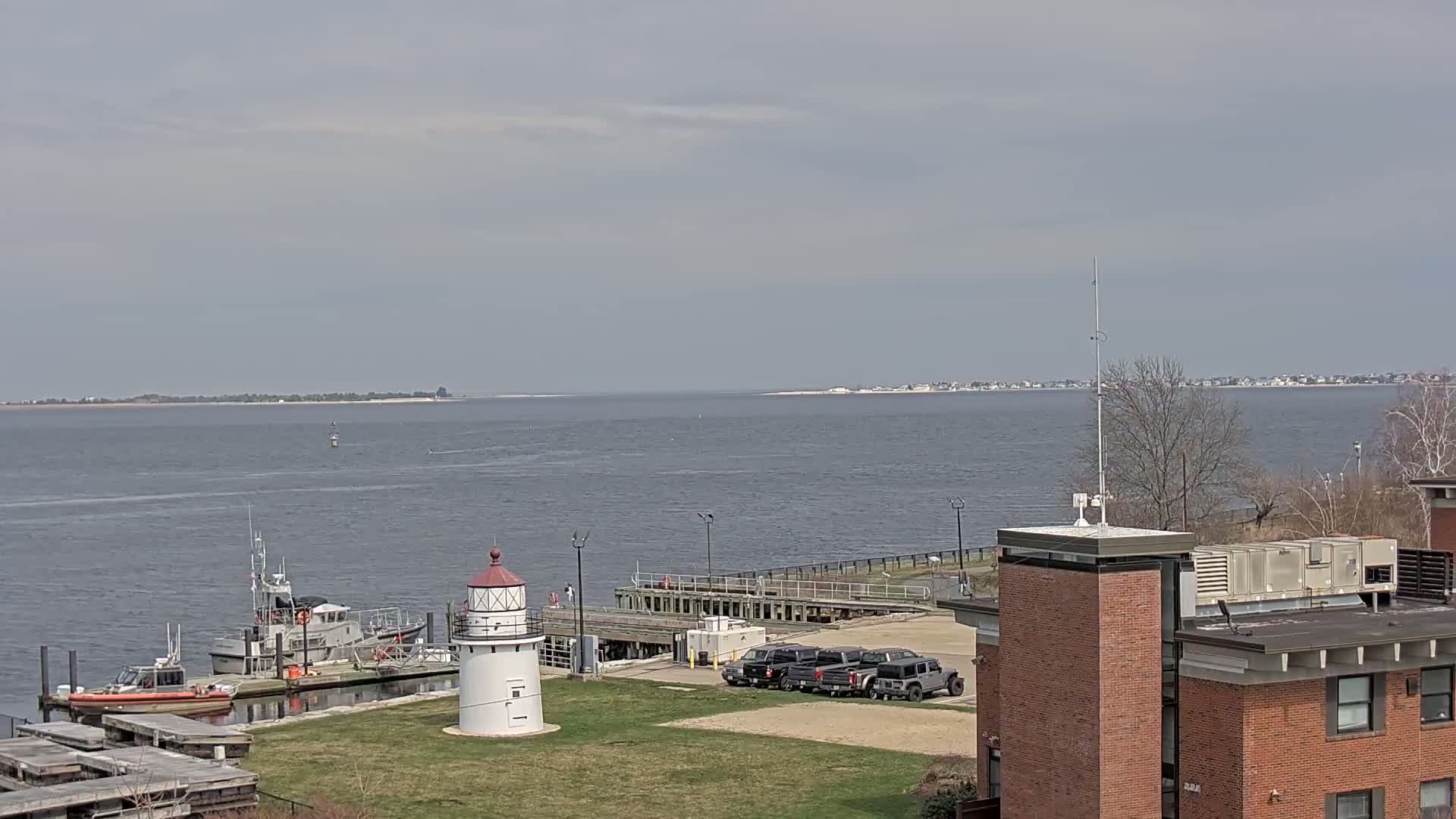 A coastal scene on an overcast day shows a lighthouse, several boats docked at a pier, and a building with a flat roof, all overlooking a calm body of water with land visible in the distance.
