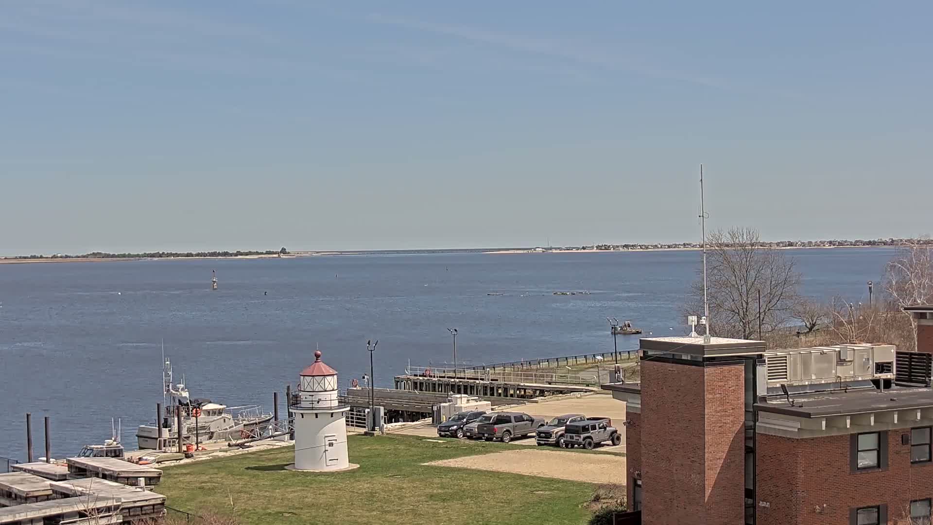 A small lighthouse stands on a grassy area next to a body of water on a sunny day, with several vehicles parked nearby, boats docked at a pier, and a building in the foreground.
