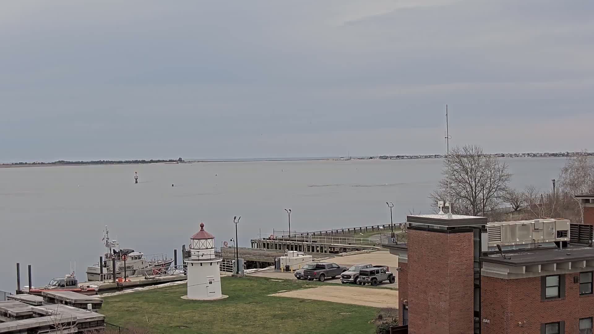 A small lighthouse stands near a dock with several vehicles parked nearby, overlooking a calm body of water under an overcast sky.
