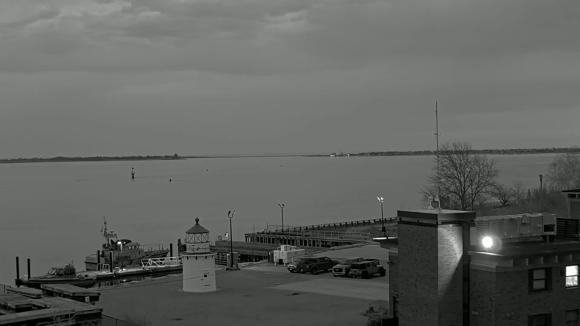 A grayscale image shows a calm body of water with a lighthouse, docked boats, and a few vehicles parked in a lot next to a building under an overcast sky.