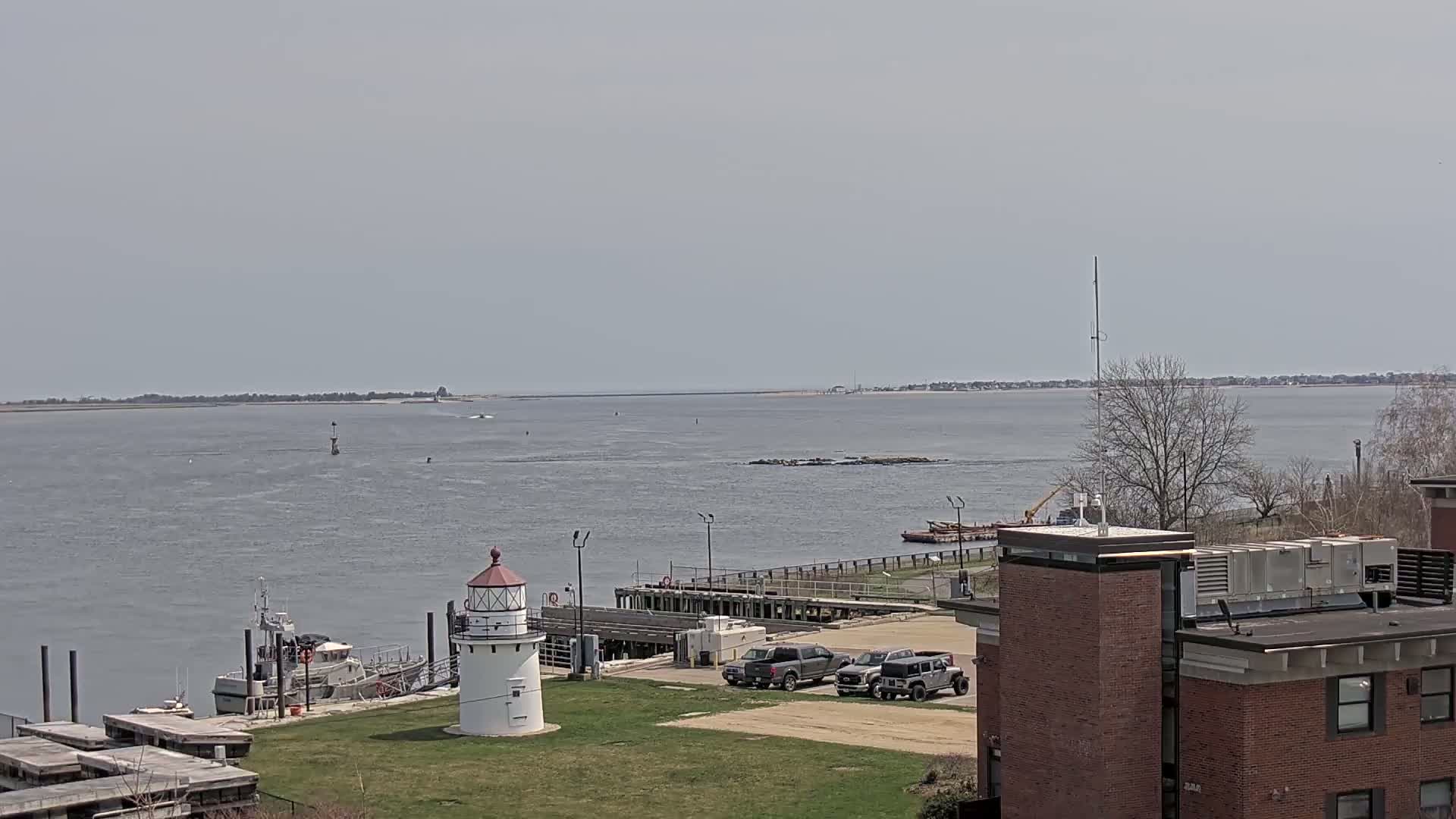 A calm, gray day overlooks a harbor with a lighthouse, docked boats, and several vehicles parked near a brick building.