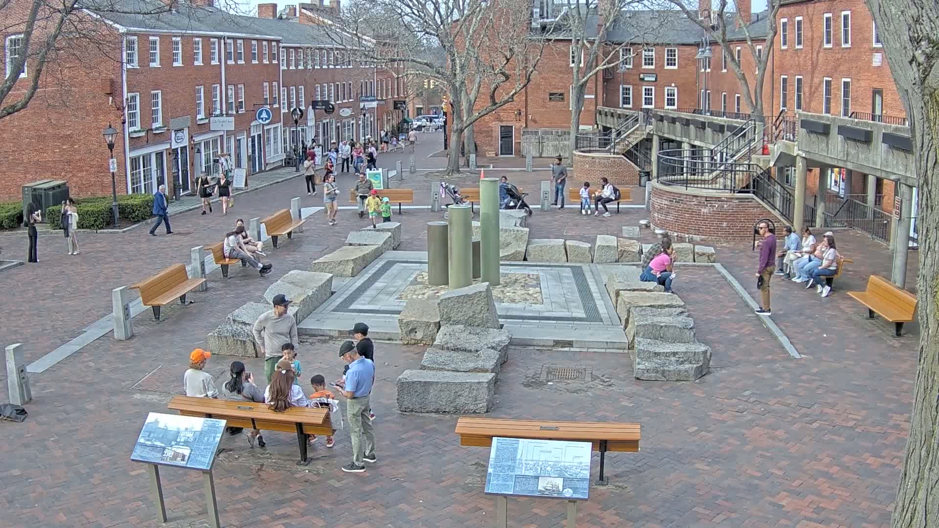 A brick town square, on a partly sunny day, features several people sitting on benches and walking around a central stone fountain, with buildings lining the square's edges.