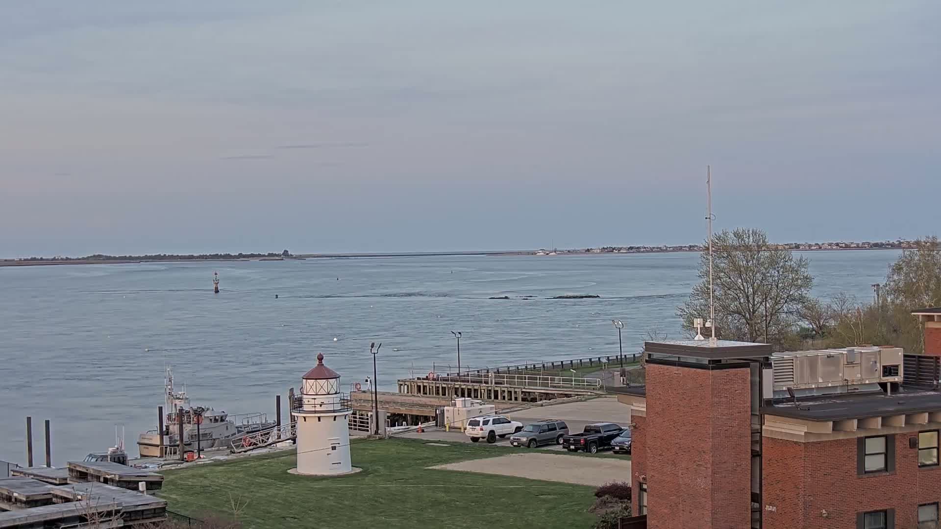 A small lighthouse stands on a grassy area near a body of water, with a boat docked nearby, under a partly cloudy sky.