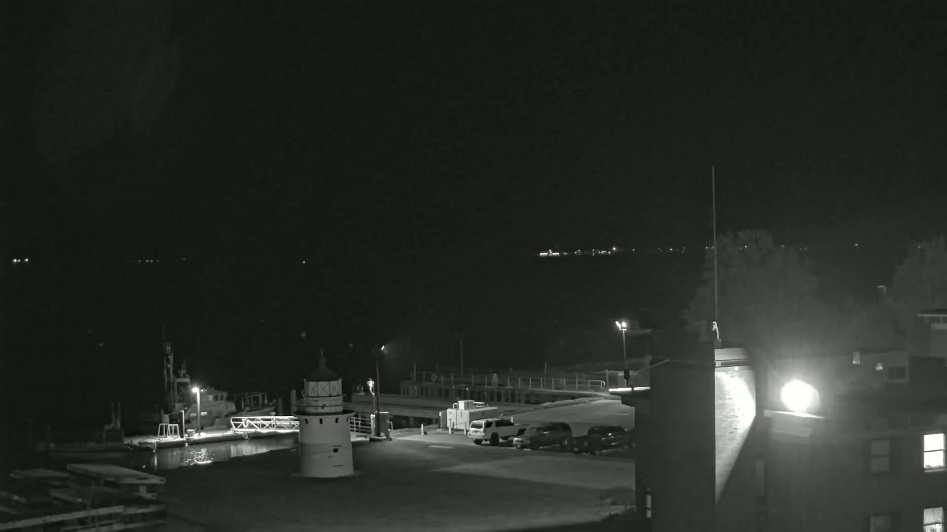 A nighttime view of a harbor with a lighthouse, docked boats, and a few parked cars under a dark sky.