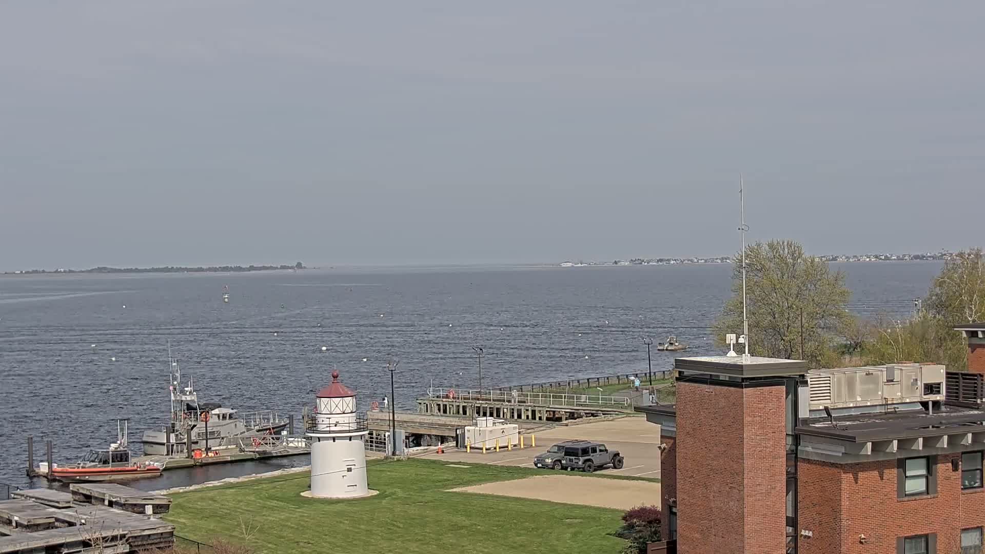A calm, gray day reveals a harbor scene with a lighthouse, docked boats, and a distant shoreline.
