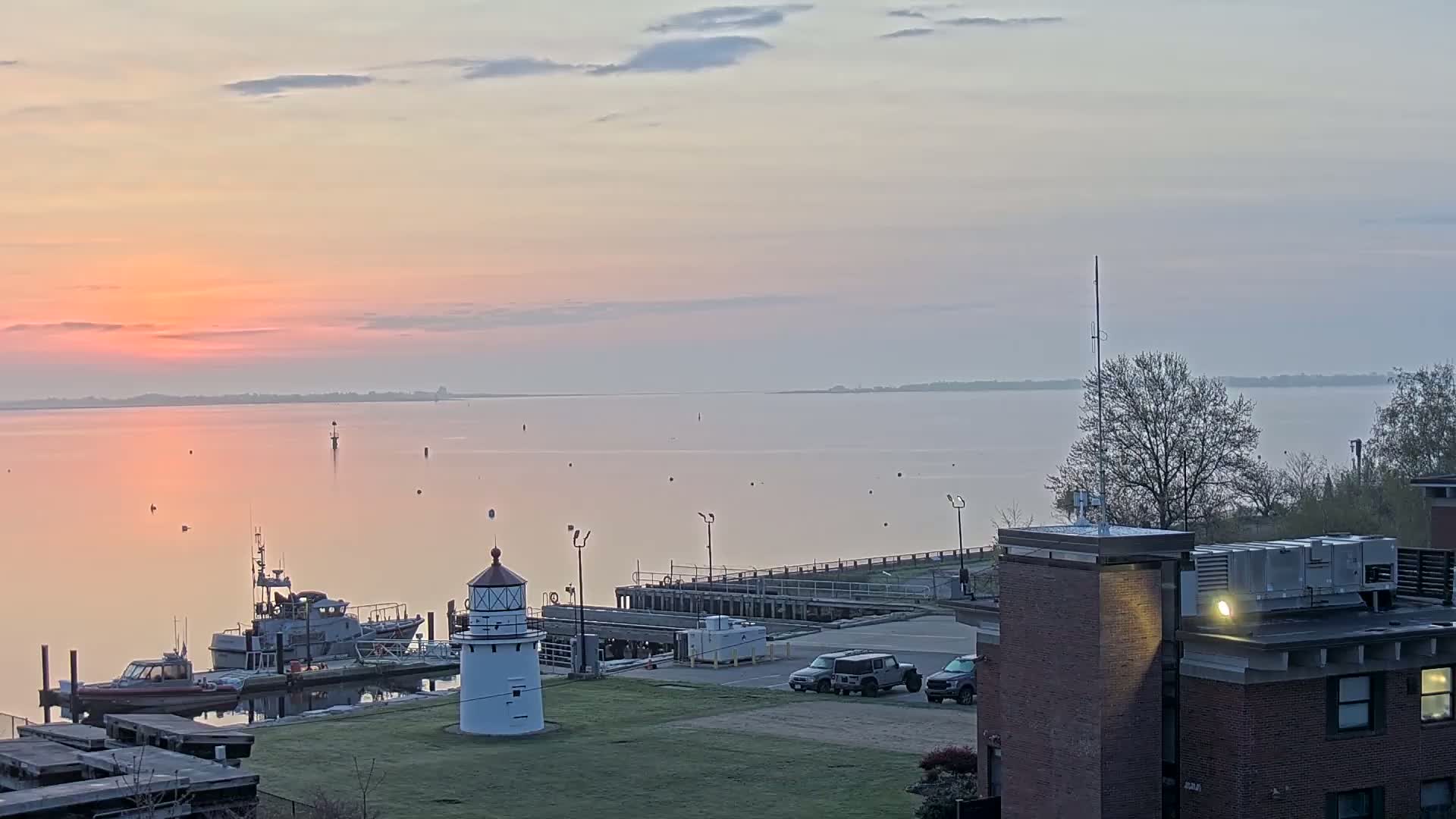 A calm, pink-hued sunrise over a body of water, with a small lighthouse, docked boats, and a building visible in the foreground.