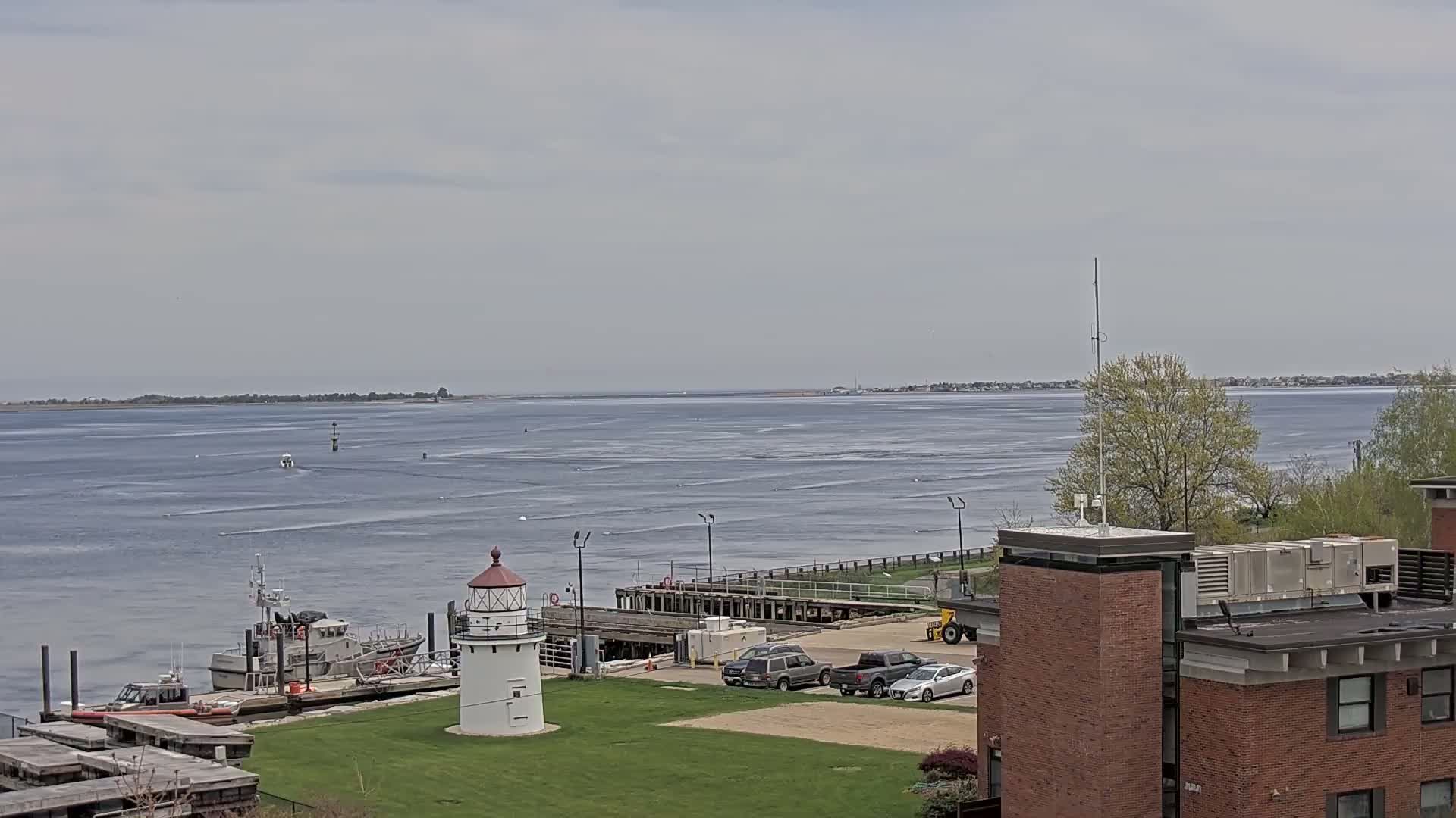 A small white lighthouse sits on a grassy area overlooking a calm body of water with several boats docked nearby, under a cloudy sky.