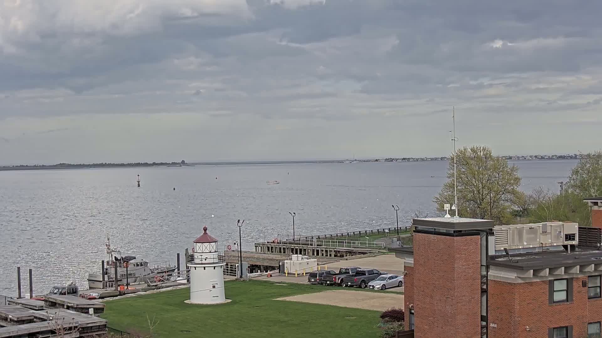 A small lighthouse sits on a grassy area next to a dock with a few boats and cars parked nearby, overlooking a calm body of water under a cloudy sky.