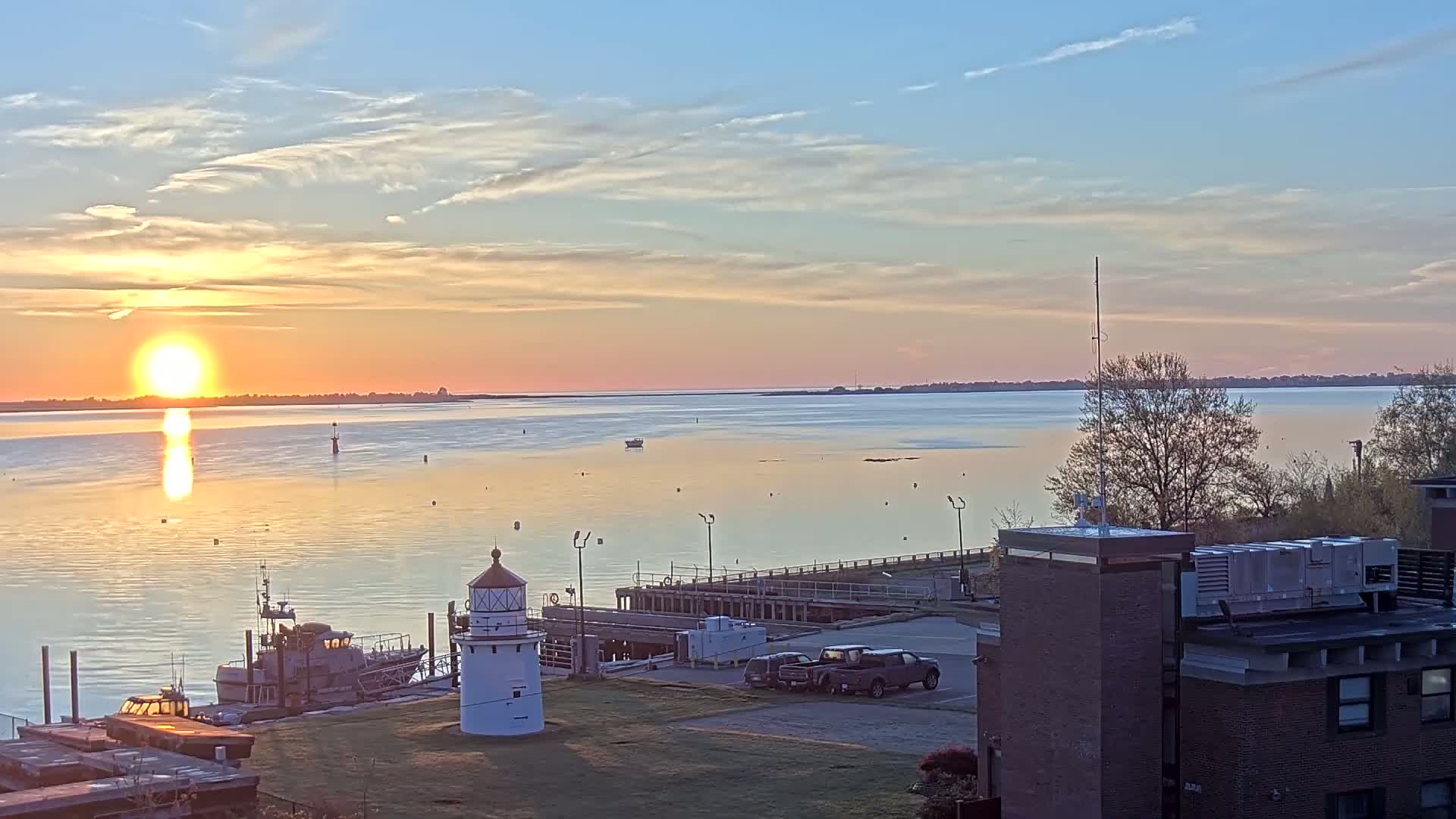 A calm body of water reflects the setting sun during a colorful sunrise, with a lighthouse, boats, and a building visible in the foreground.