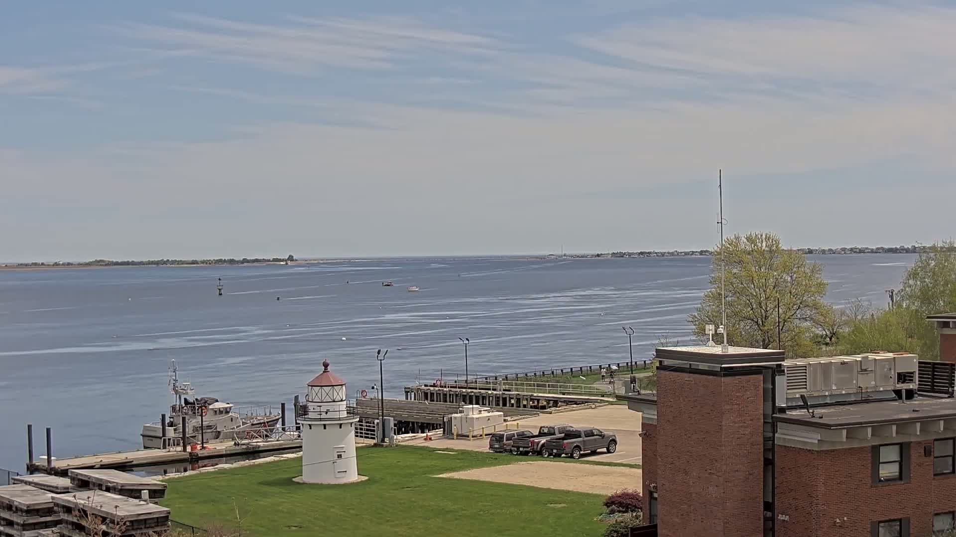 A small lighthouse stands on a grassy area next to a dock with a boat, overlooking a calm body of water under a mostly sunny sky.