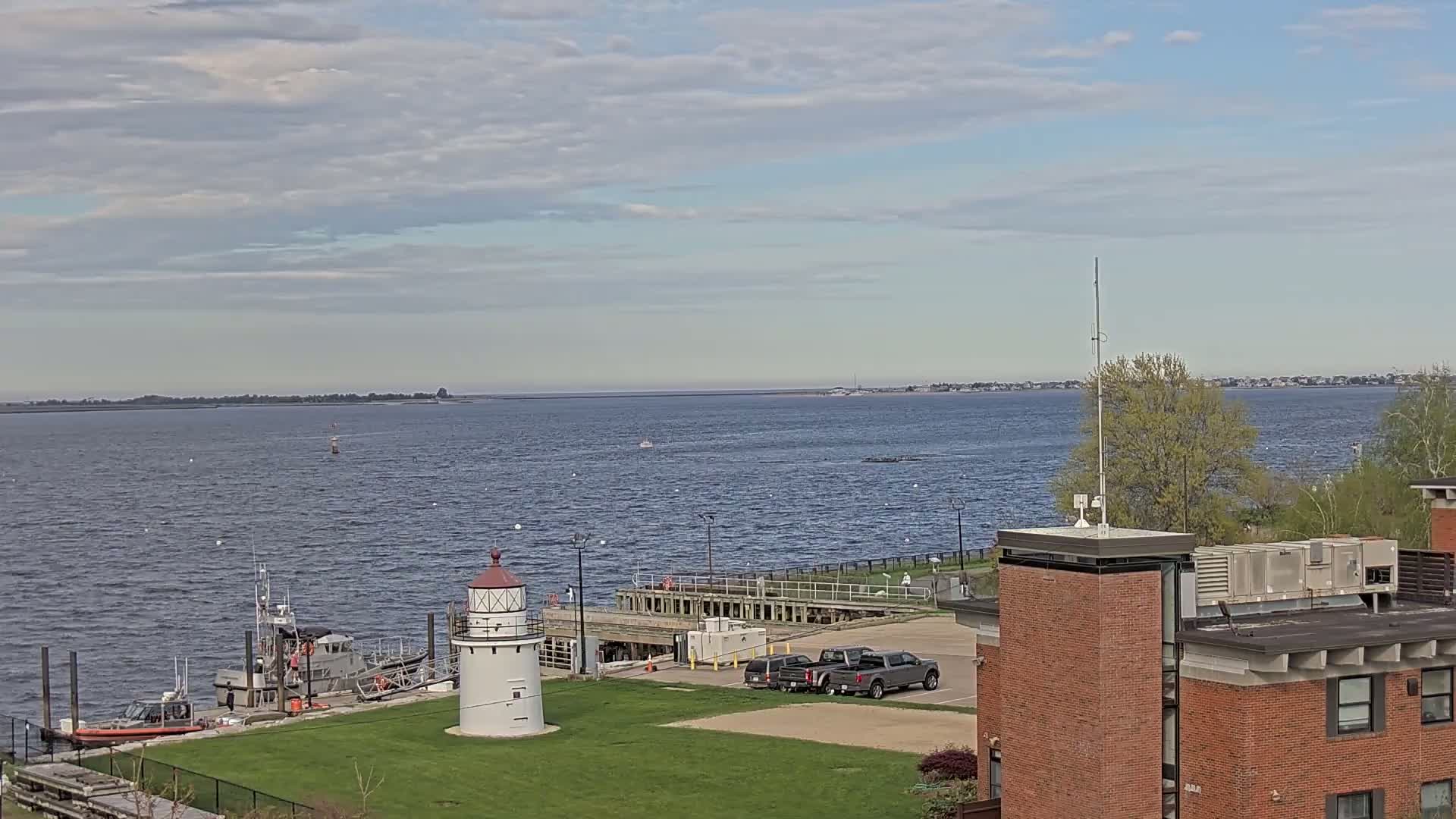 A white lighthouse stands on a grassy area next to a body of water under a partly cloudy sky, with several boats and vehicles visible nearby.