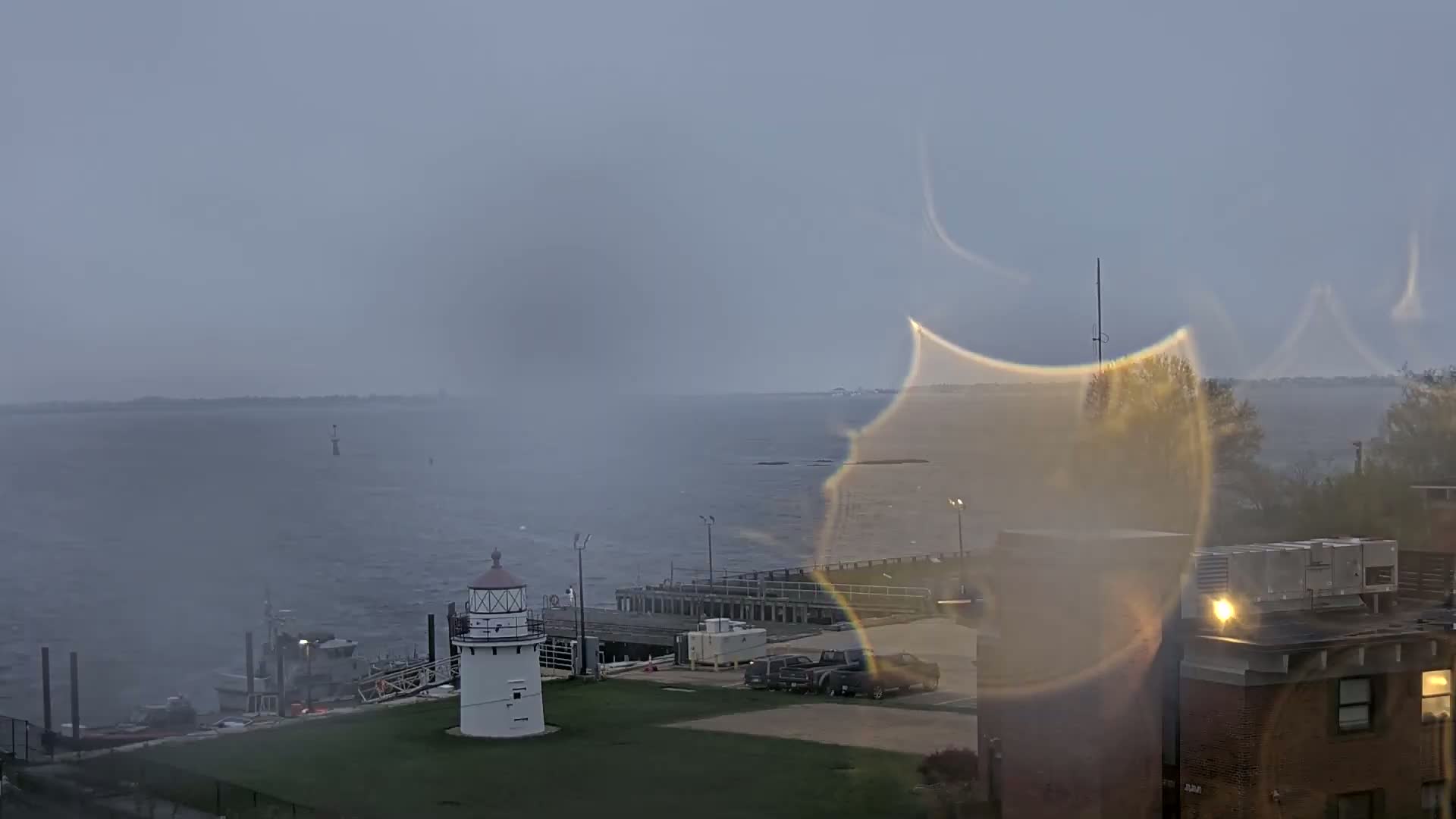 A view through a rain-streaked window shows a lighthouse on a grassy area near a dock, a body of water with a boat, and a building in the foreground under an overcast sky.