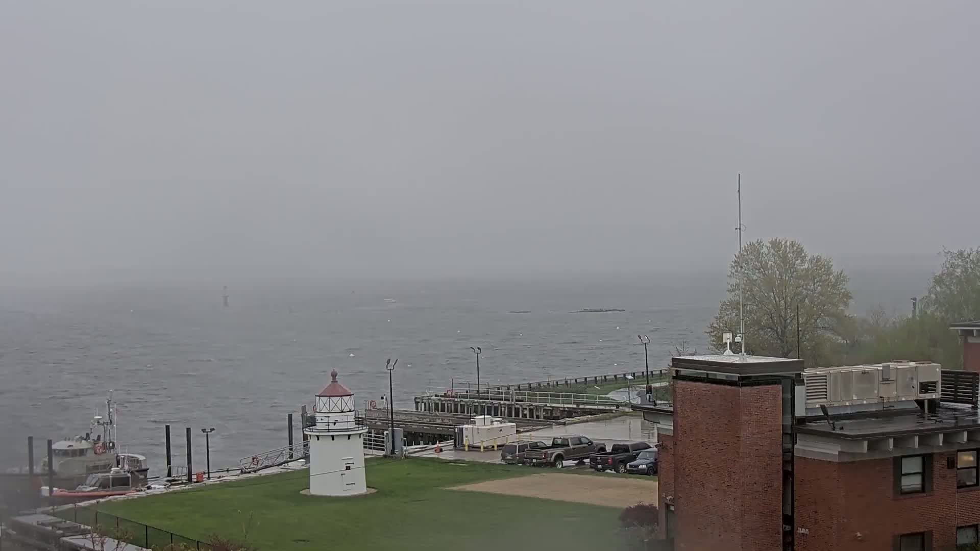 A small lighthouse stands on a grassy area overlooking a choppy sea under a foggy, overcast sky, with a Coast Guard boat moored nearby and a building visible in the lower right corner.