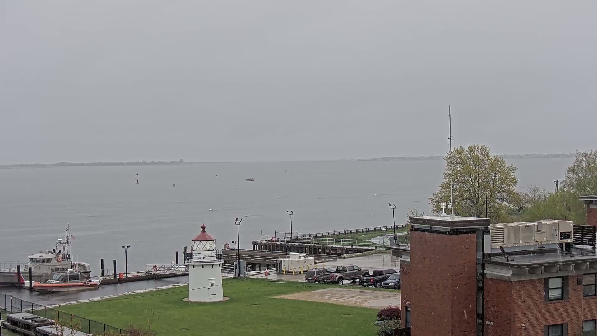 A lighthouse stands on a grassy area next to a harbor with a Coast Guard boat and other vessels, under an overcast sky with light snow or rain falling.