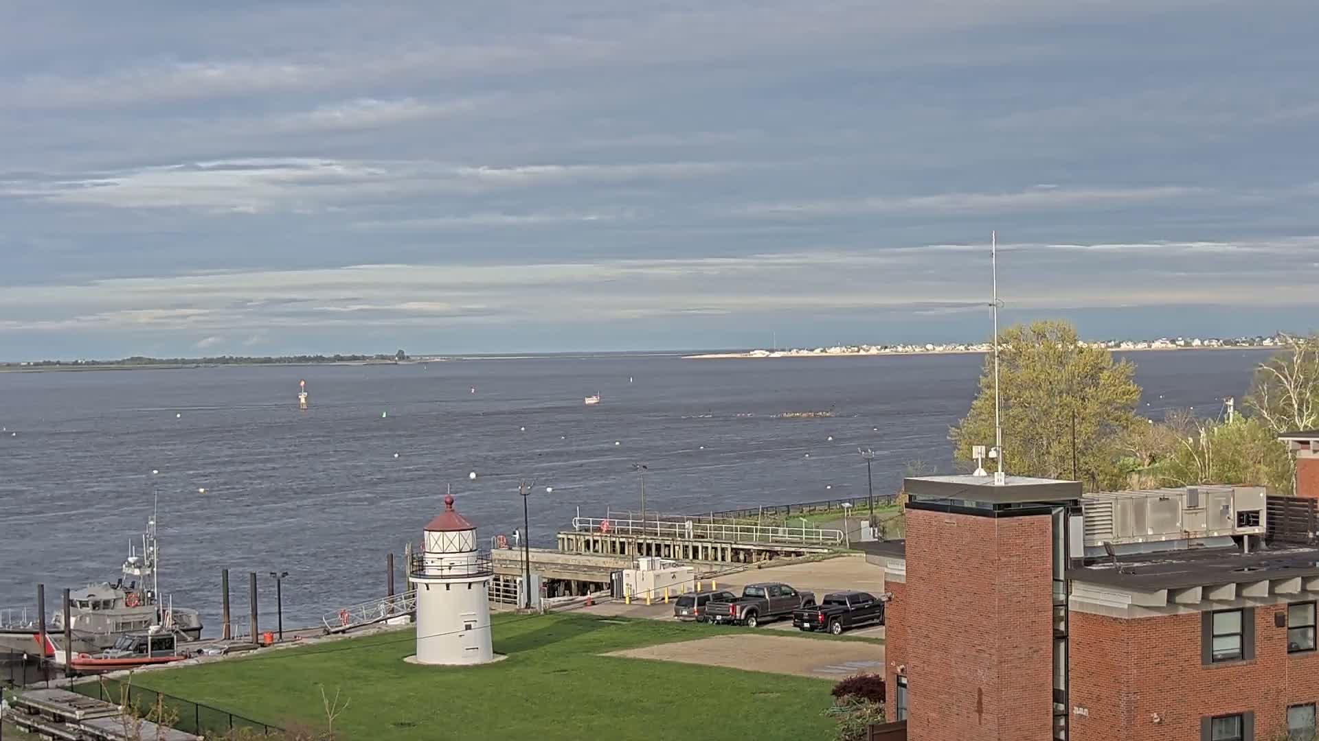 A small lighthouse stands on a grassy area next to a Coast Guard boat, overlooking a calm body of water under a partly cloudy sky, with a distant shoreline visible.
