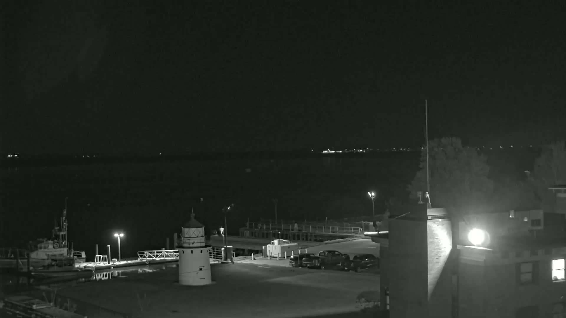 A nighttime, dark gray-scale image shows a harbor with a lighthouse, several docked boats, and a few parked vehicles under a clear, dark sky.