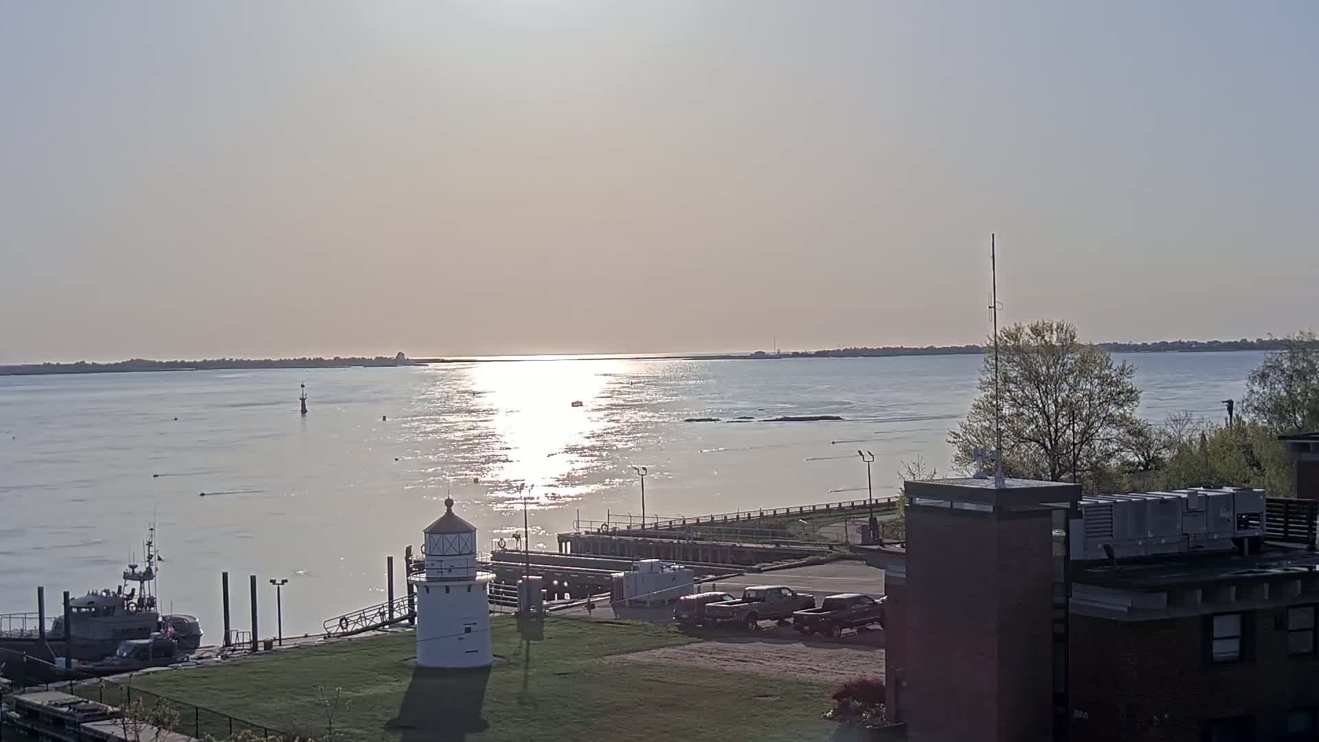 A white lighthouse stands on a grassy area next to a calm river reflecting sunlight, with a small boat docked nearby and a larger vessel further out on the water under a bright, clear sky.