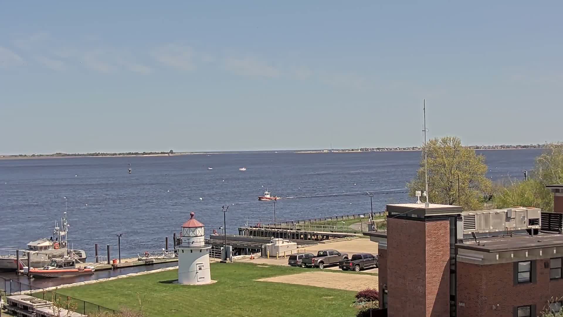 A coast guard boat is on the water near a lighthouse and docked boats, under a clear blue sky.