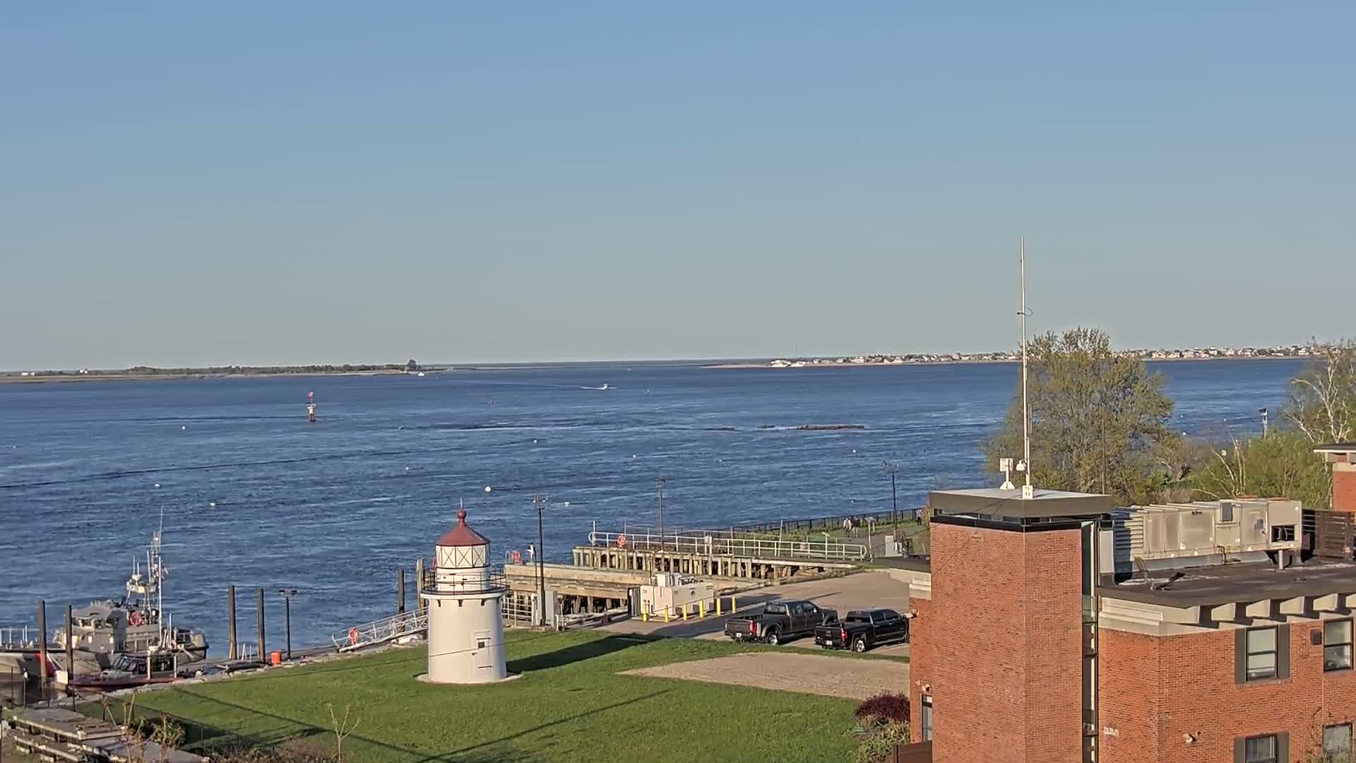 A small lighthouse stands near a Coast Guard vessel by a calm body of water under a clear blue sky, with a distant shoreline visible.