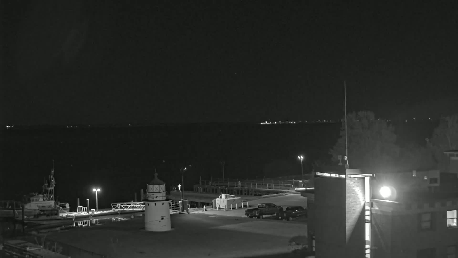 A nighttime view of a harbor with a lighthouse, docked boats, and a building, under a clear, dark sky.