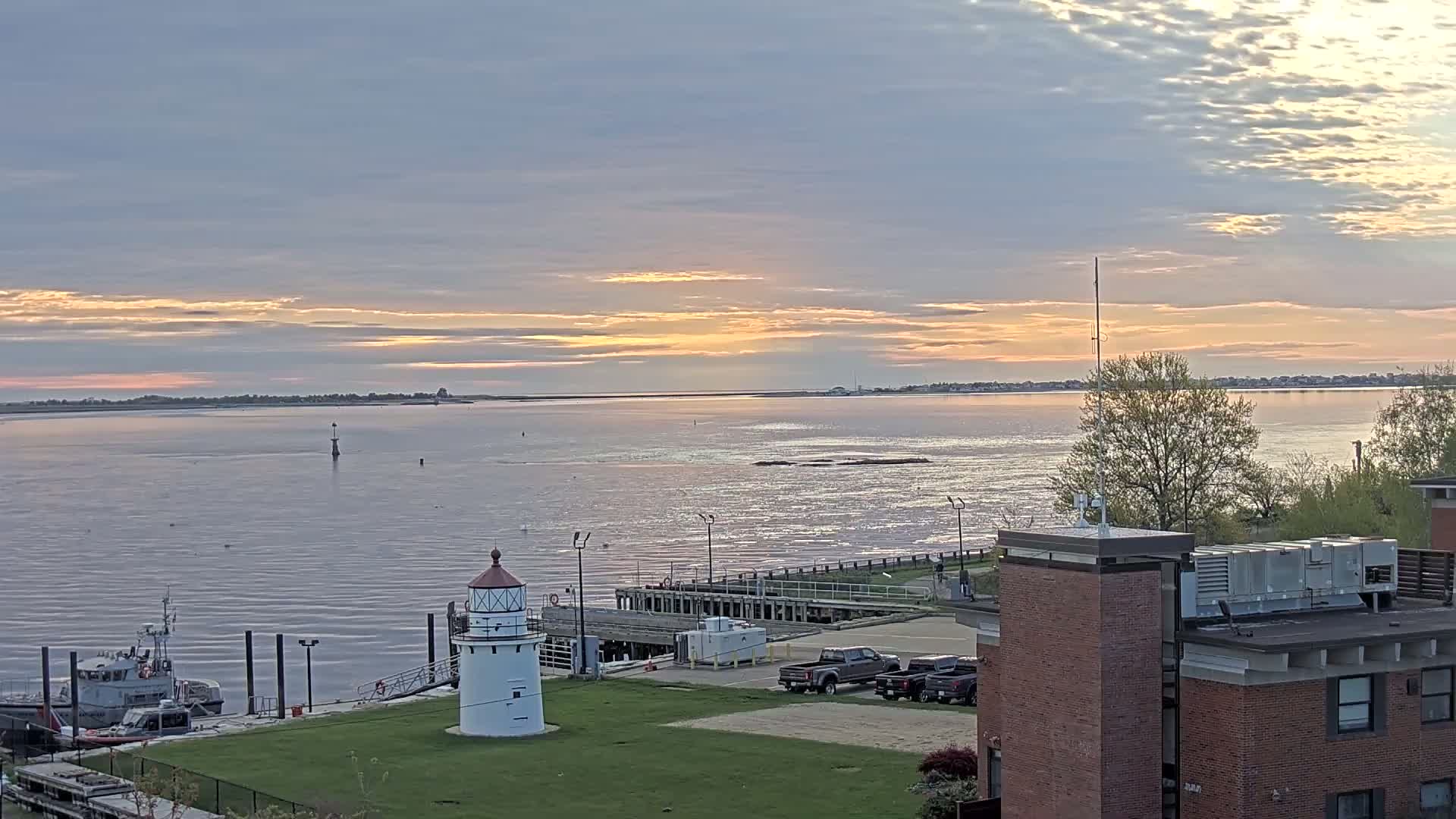 A coastal scene at sunrise shows a lighthouse, a small harbor with a boat, and calm water reflecting the light of the sky under a partly cloudy sky.