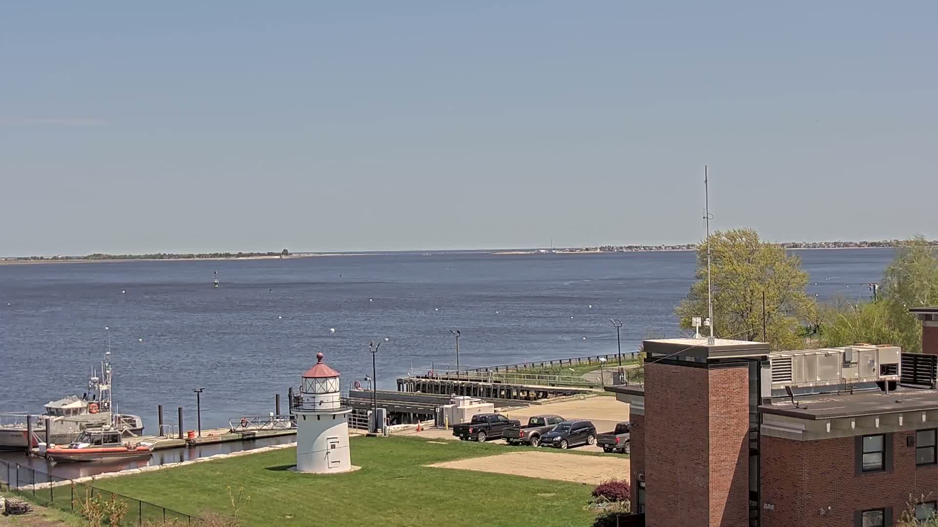 A coastal scene on a sunny day shows a lighthouse, docked boats, and several vehicles parked in a lot near a brick building, all overlooking a calm body of water with a distant shoreline.