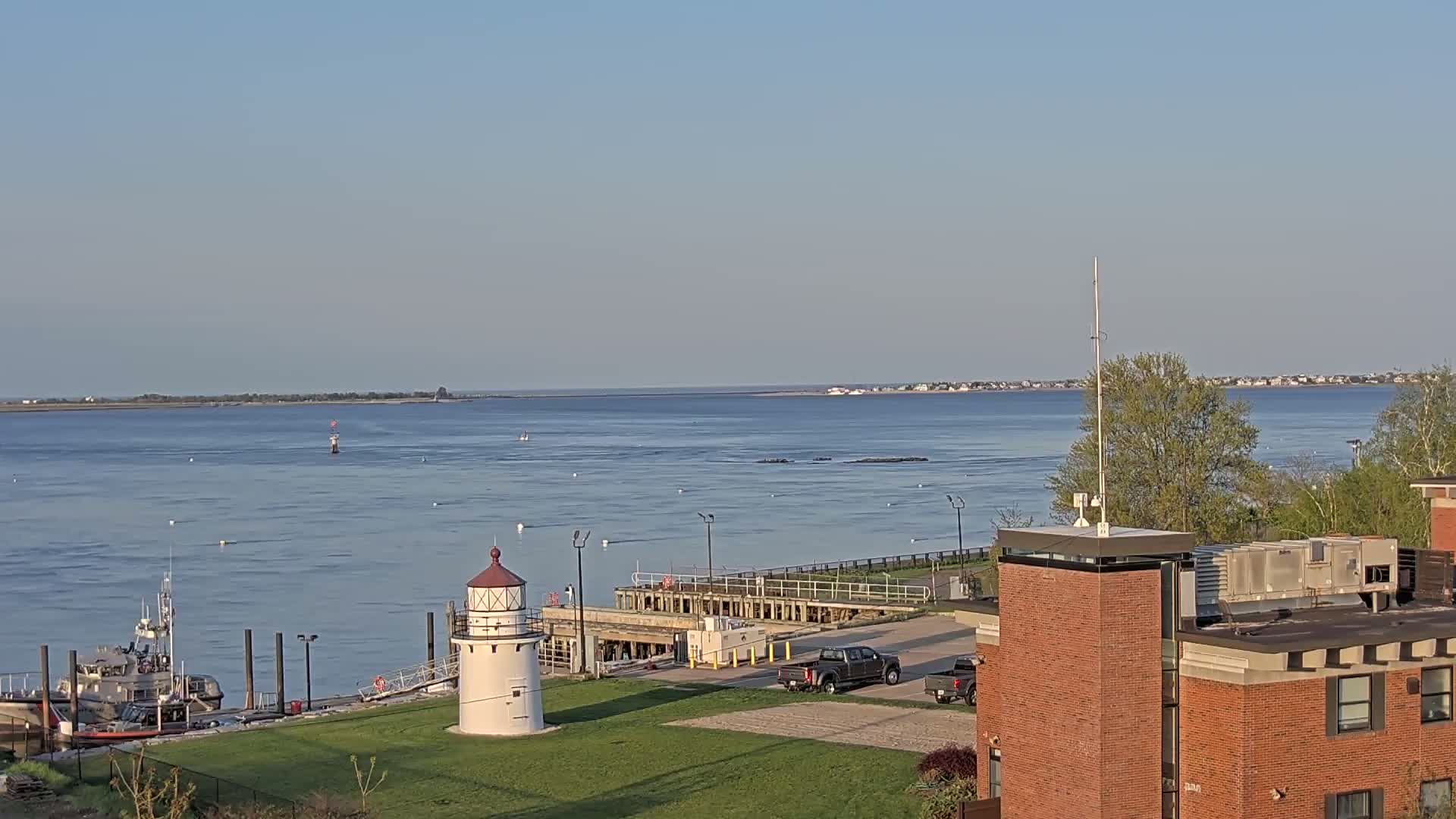 A calm, wide body of water is viewed from a high vantage point, showing a small lighthouse, a docked boat, and a distant shoreline under a clear sky.