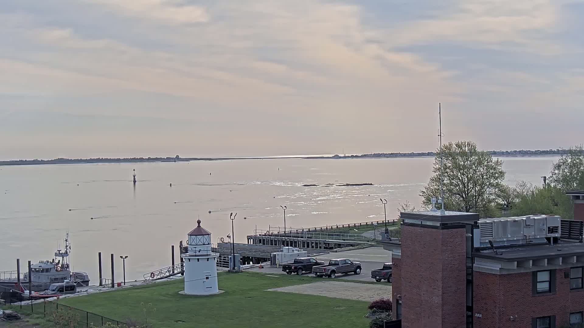 A small lighthouse sits on a grassy area near a dock and calm body of water under a mostly cloudy sky.