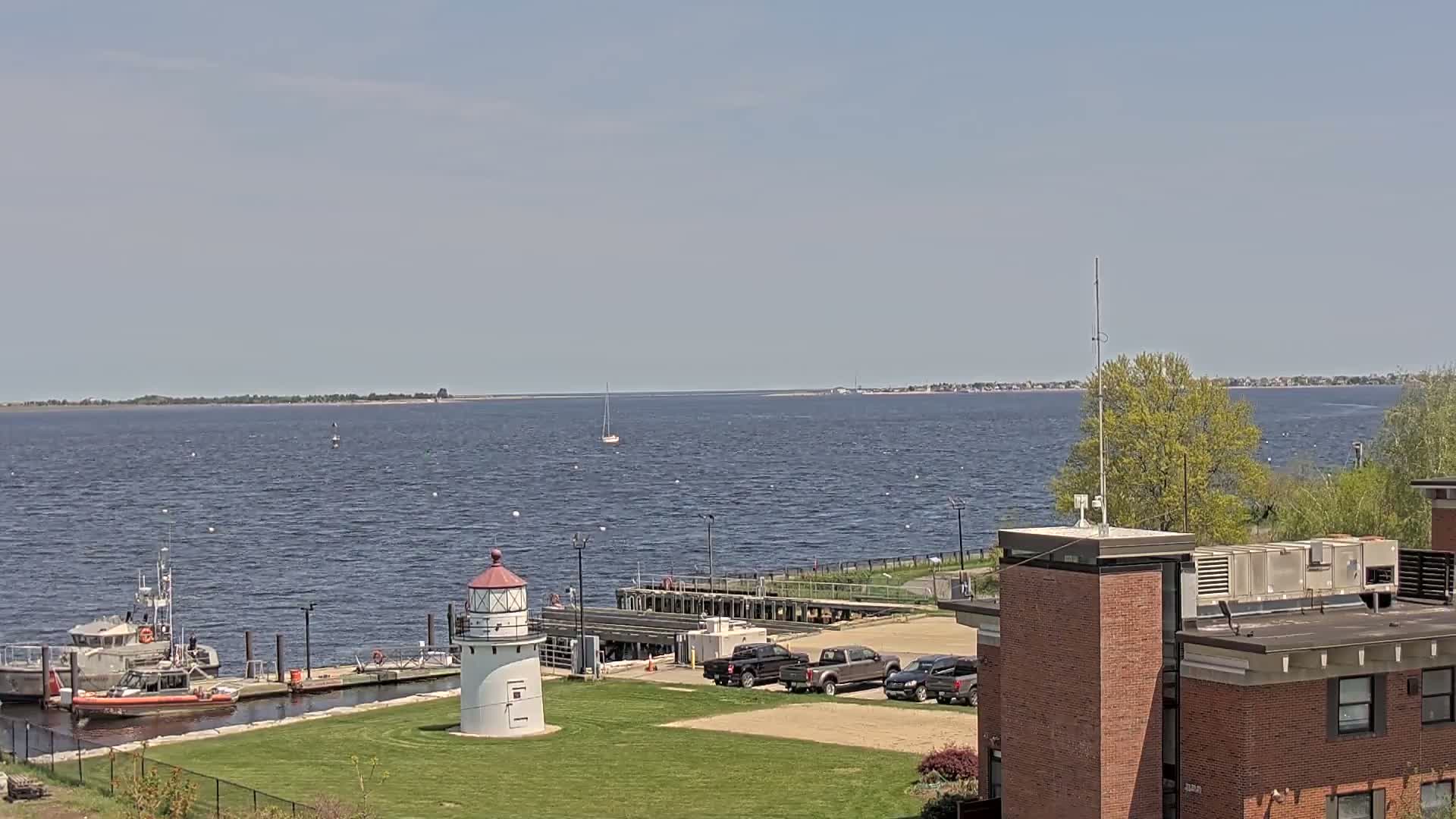 A coastal scene on a sunny day shows a body of water with a sailboat, a Coast Guard vessel at a dock near a lighthouse, several trucks parked nearby, and a brick building in the foreground.