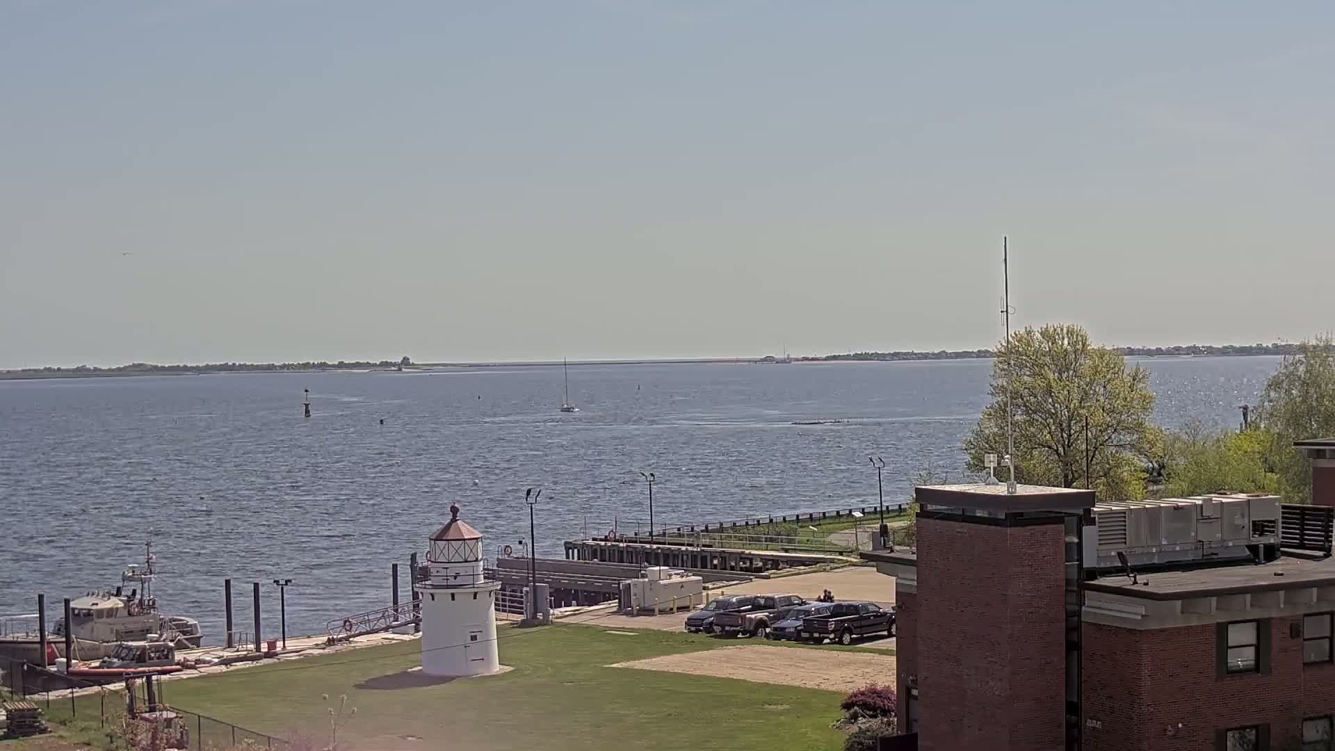 A small lighthouse stands near a Coast Guard boat by a calm body of water under a clear blue sky, with several cars parked nearby and a distant shoreline visible.