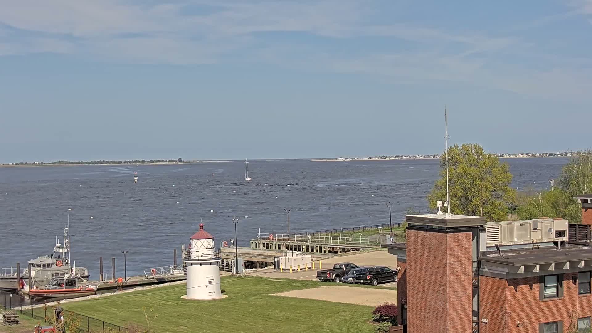 A coast guard boat is docked near a small lighthouse and a larger building on a partly sunny day overlooking a wide body of water.