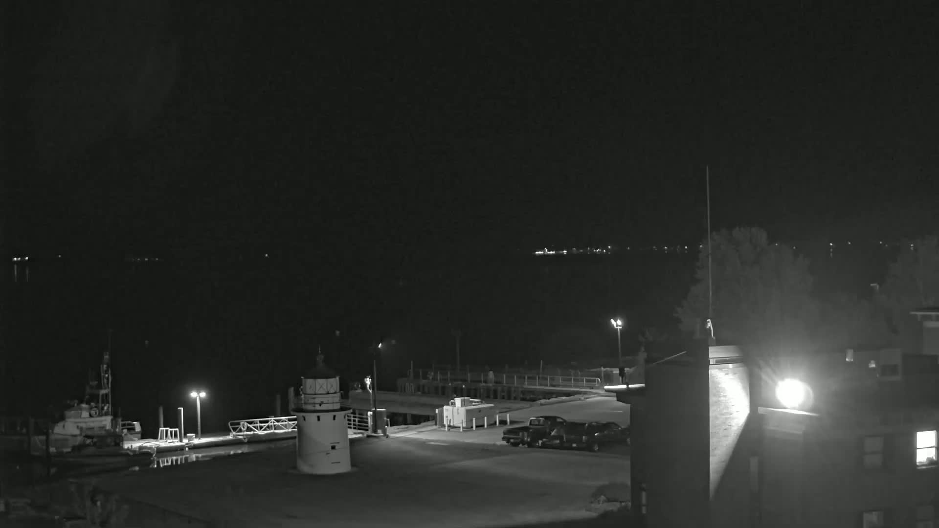 A nighttime, black-and-white view shows a small lighthouse and a docked boat near a building with cars parked in the lot, all situated by a dark body of water under a clear sky.