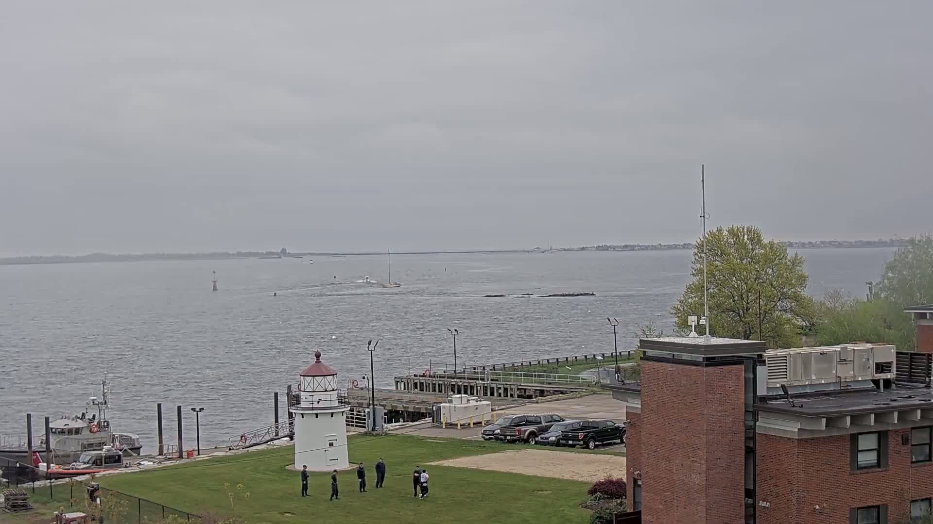 A coast guard boat is docked near a lighthouse on a grassy area next to a brick building, with several people standing on the grass and a sailboat visible in the overcast water.