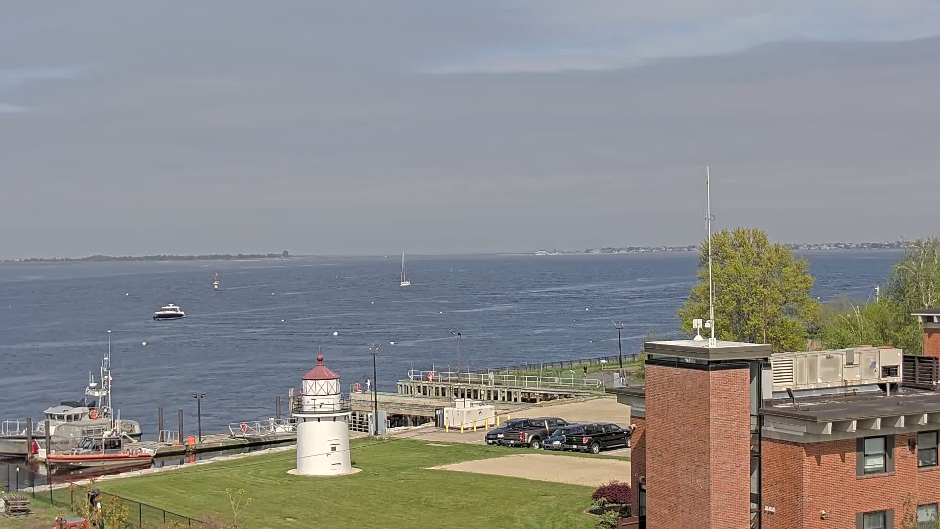 A small lighthouse stands on a grassy area near a harbor with several boats, including a Coast Guard vessel, under a mostly cloudy sky.