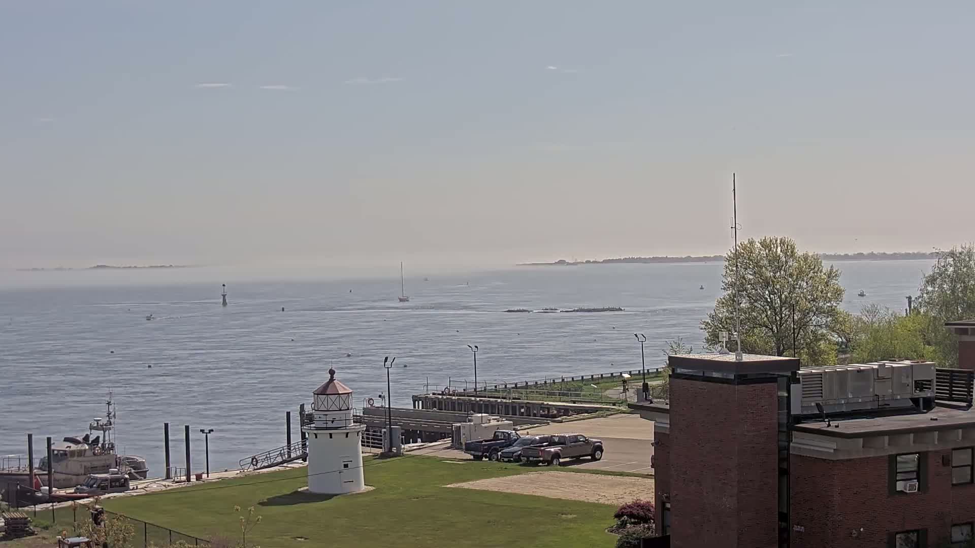 A small lighthouse sits on a grassy area next to a dock and calm, hazy water with a few boats and a distant shoreline.