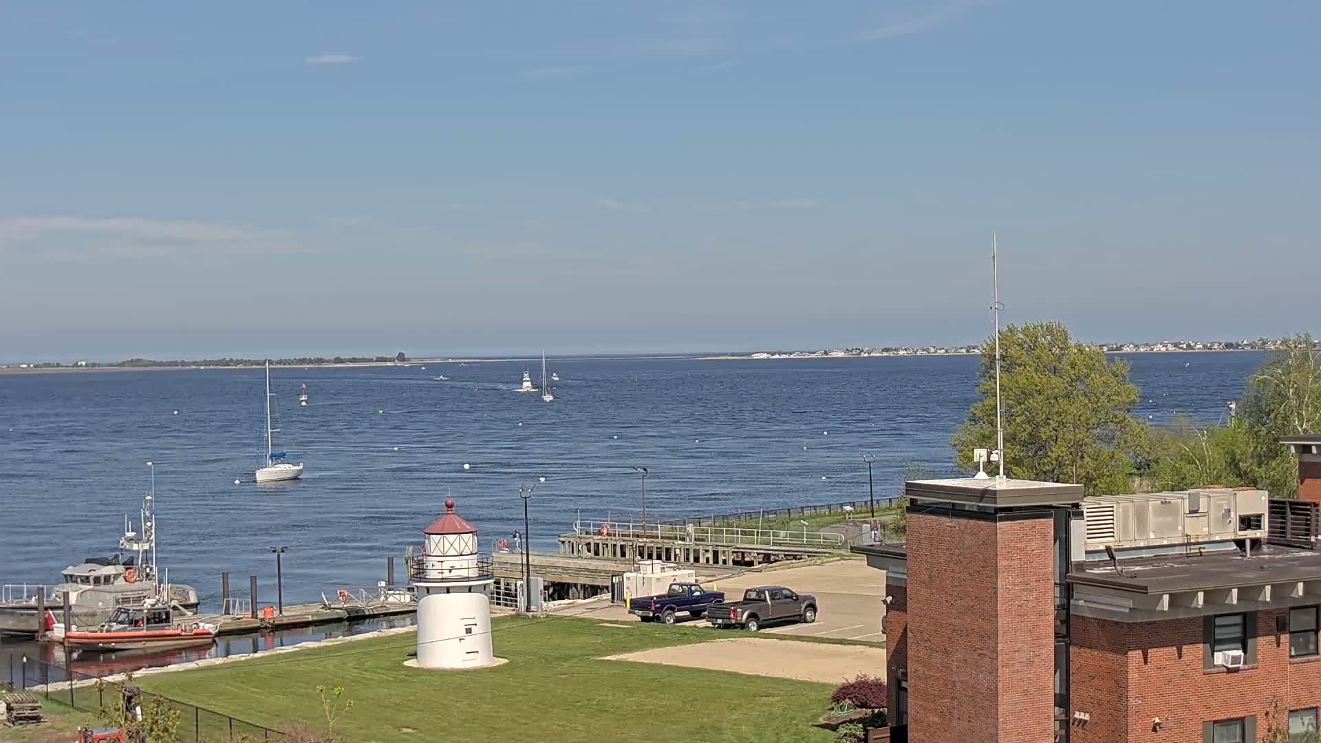 A sunny day shows a harbor with several boats, a lighthouse, and a Coast Guard vessel, all overlooking a body of water with a distant shoreline.
