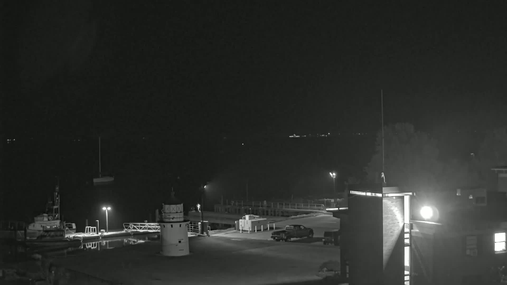 A nighttime, black and white view of a harbor with a small lighthouse, docked boats, a truck, and a building under a clear, dark sky.