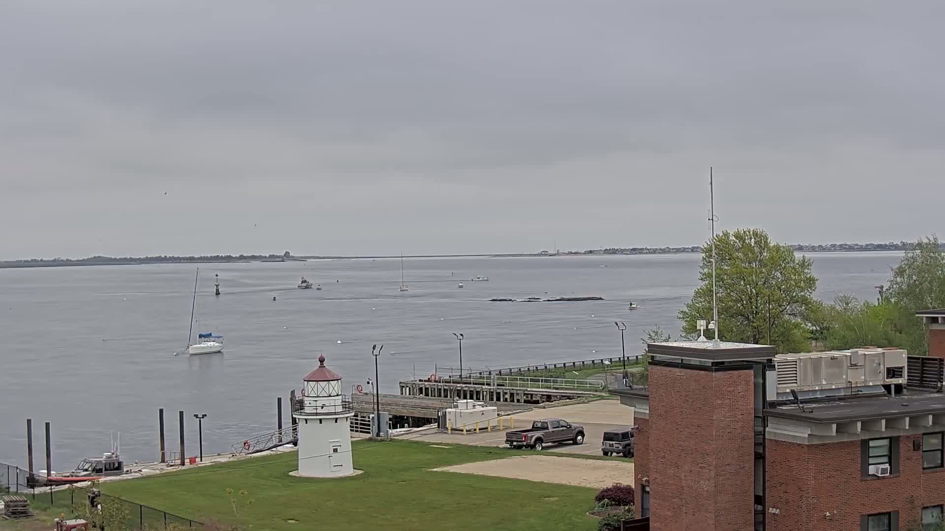 A small white lighthouse stands on a grassy area overlooking a calm body of water under an overcast sky, with several boats visible on the water and a brick building in the foreground.