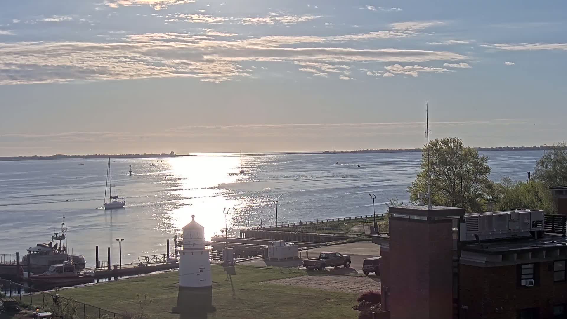 A calm, sunlit waterway with several boats and a small lighthouse near a dock and building under a partly cloudy sky.