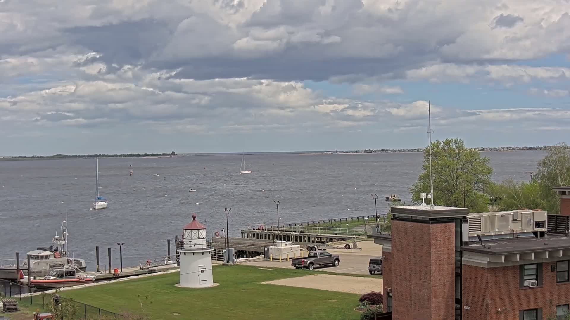 A small lighthouse stands on a grassy area next to a dock with a Coast Guard boat and other vessels, overlooking a calm body of water under a partly cloudy sky.