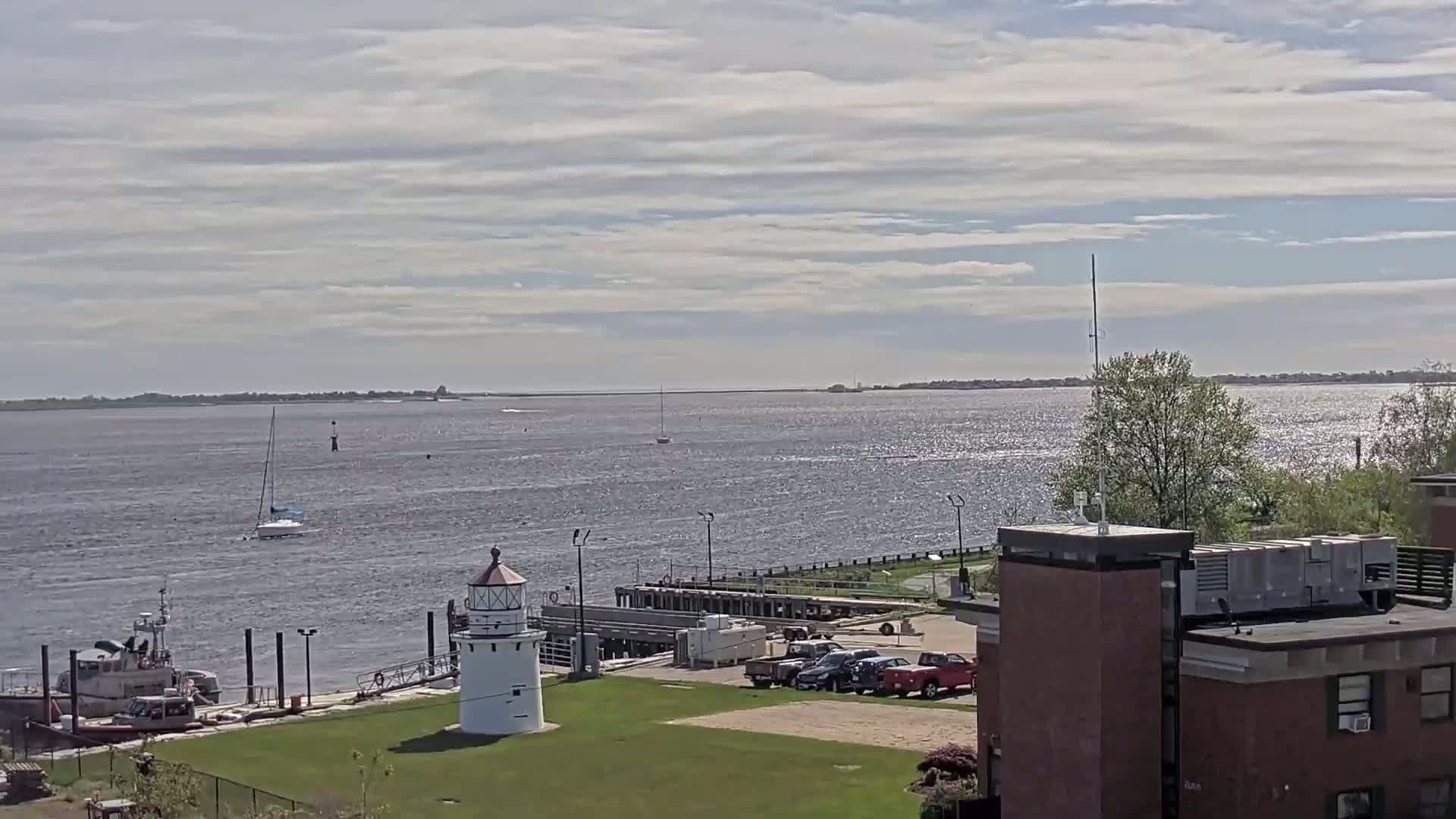 A small lighthouse stands on a grassy area next to a harbor with a few boats, a Coast Guard vessel, and several parked cars, all under a partly sunny sky.