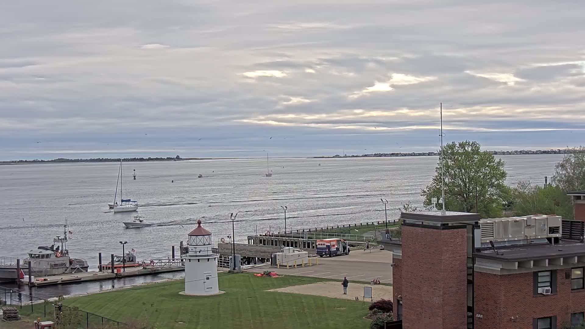 A small lighthouse sits on a grassy area next to a harbor with several boats, a coast guard vessel, and a building under a mostly cloudy sky.