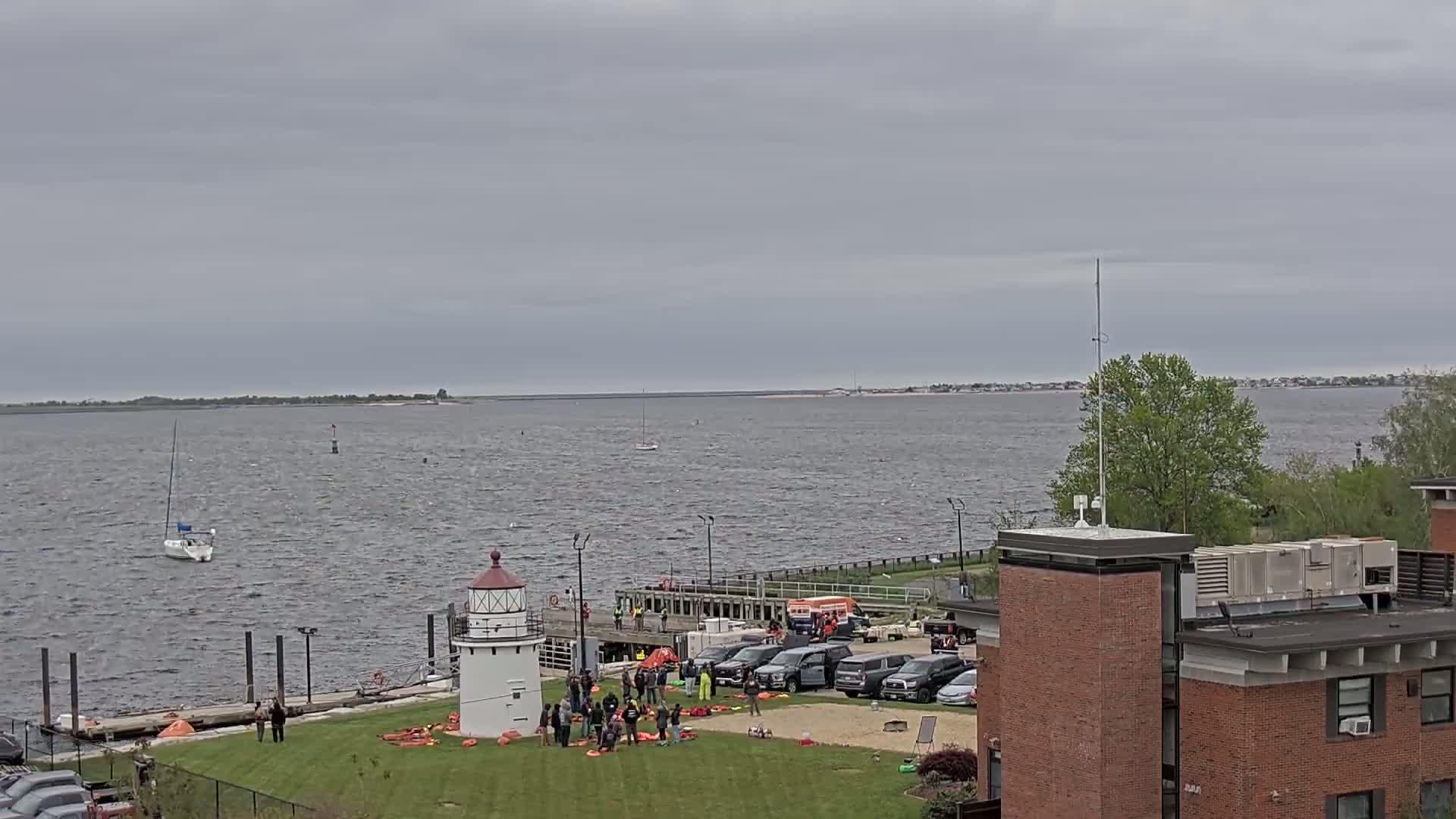 A group of people are gathered near a small lighthouse on a grassy area next to a body of water under an overcast sky; several vehicles are parked nearby.